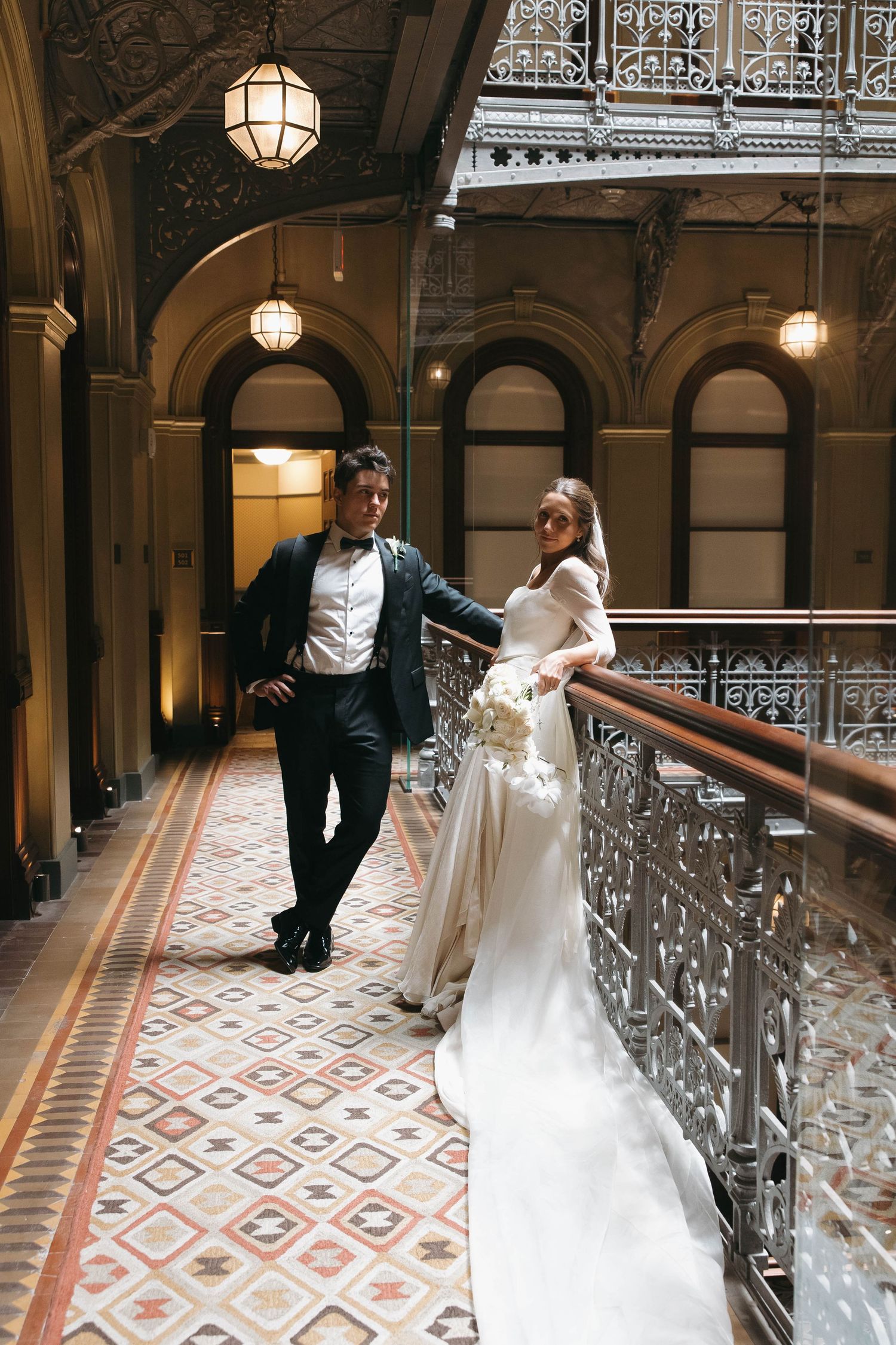 Bride in white gown and groom in dark suit pose on ornate balcony of elegant historic building with mosaic tile floors.