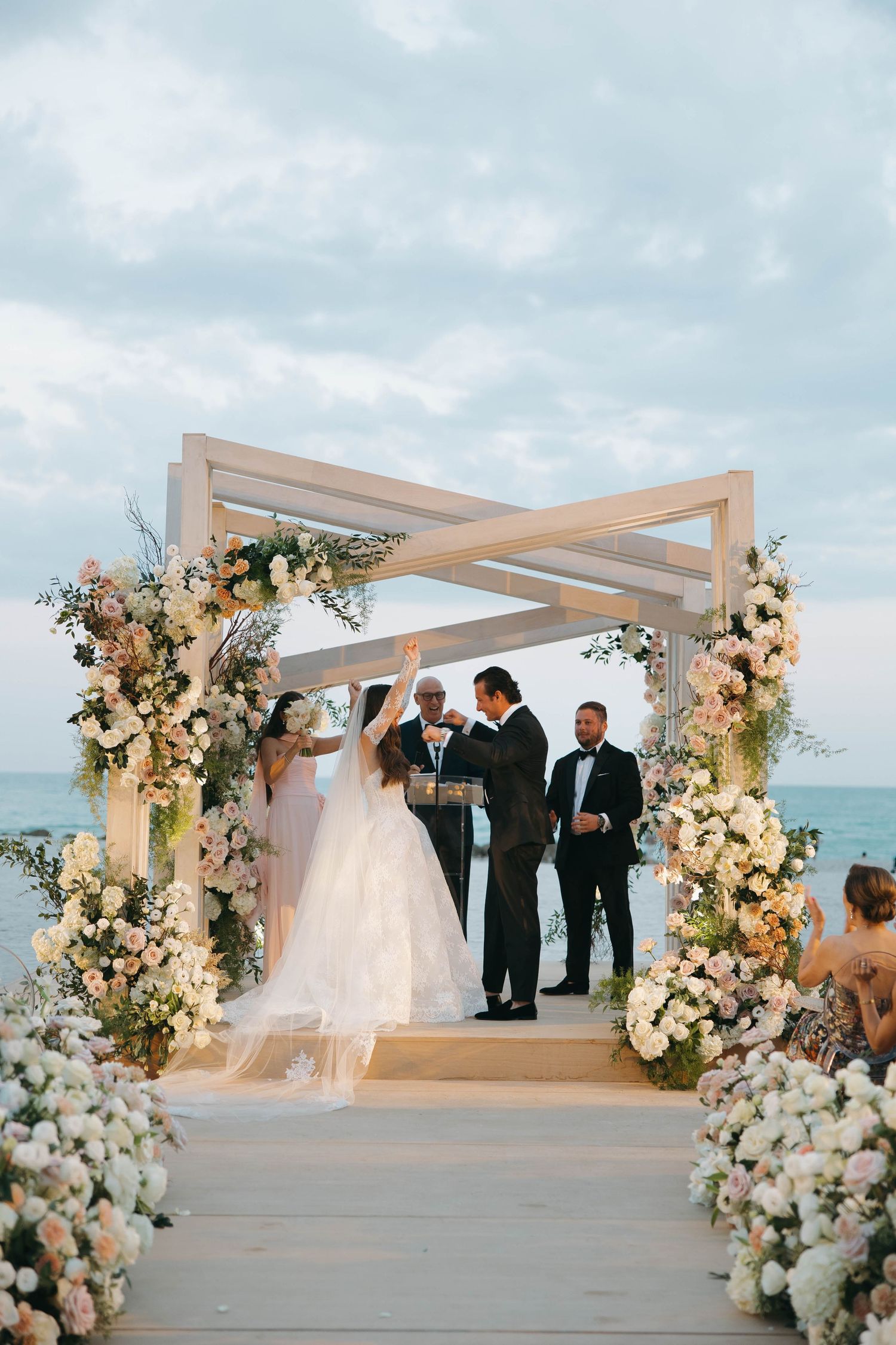 Seaside wedding ceremony under a modern white pergola decorated with luxurious white and pink flowers.