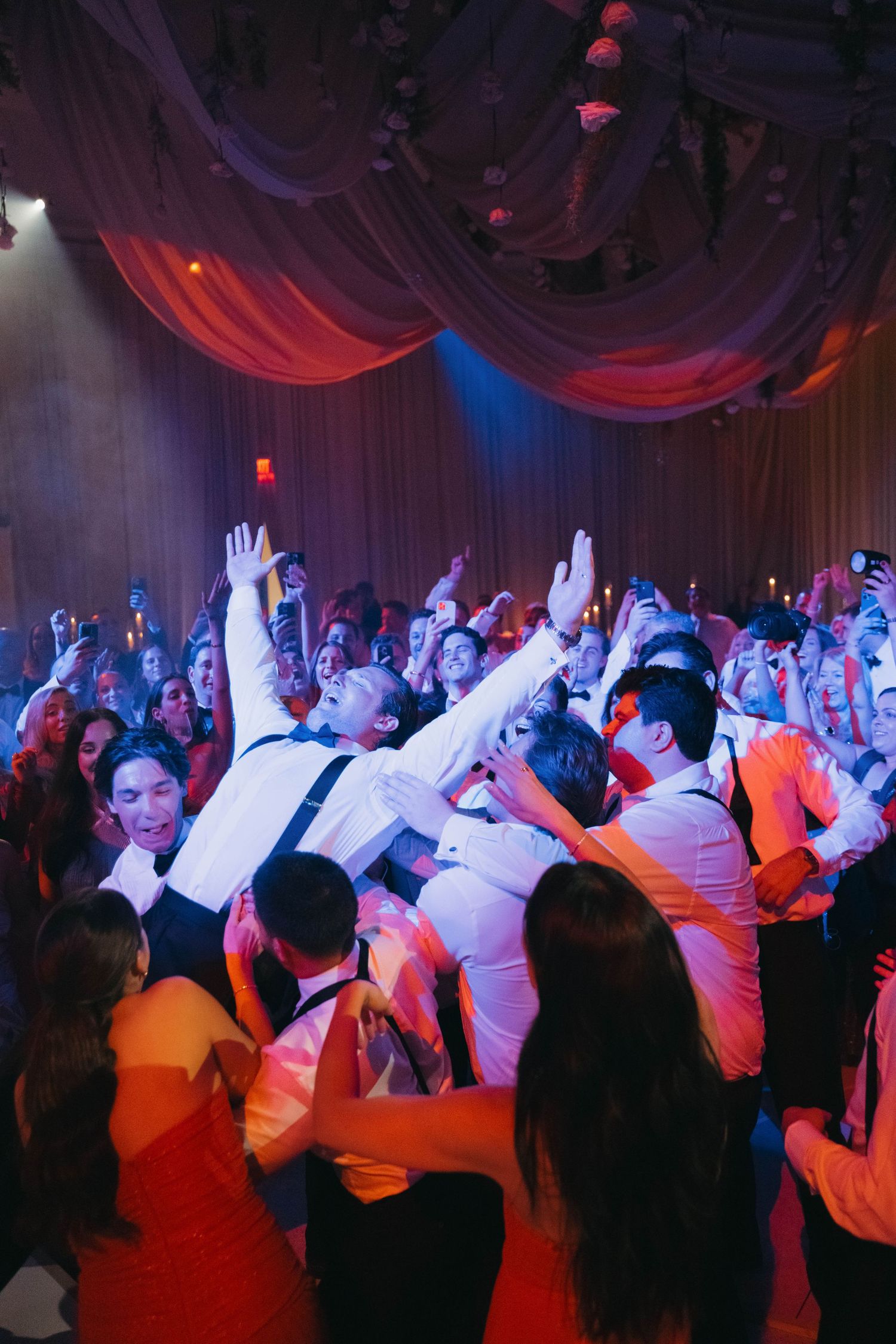 Energetic crowd with raised hands dancing at a wedding reception under colorful mood lighting.