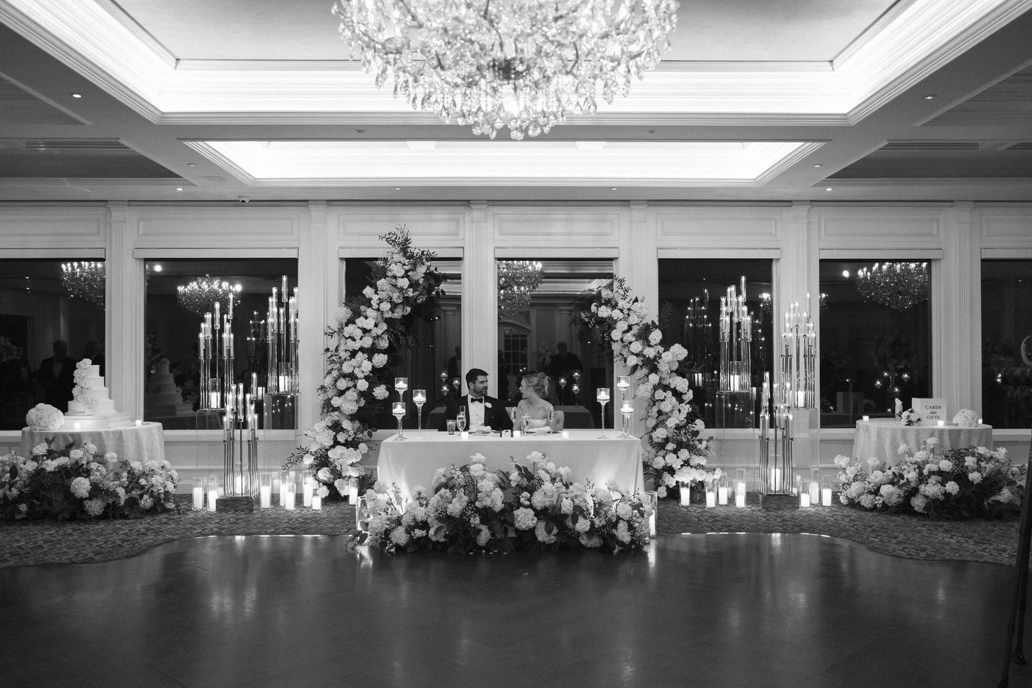 Elegant wedding reception setup with crystal chandelier and floral arrangements on white-draped tables.