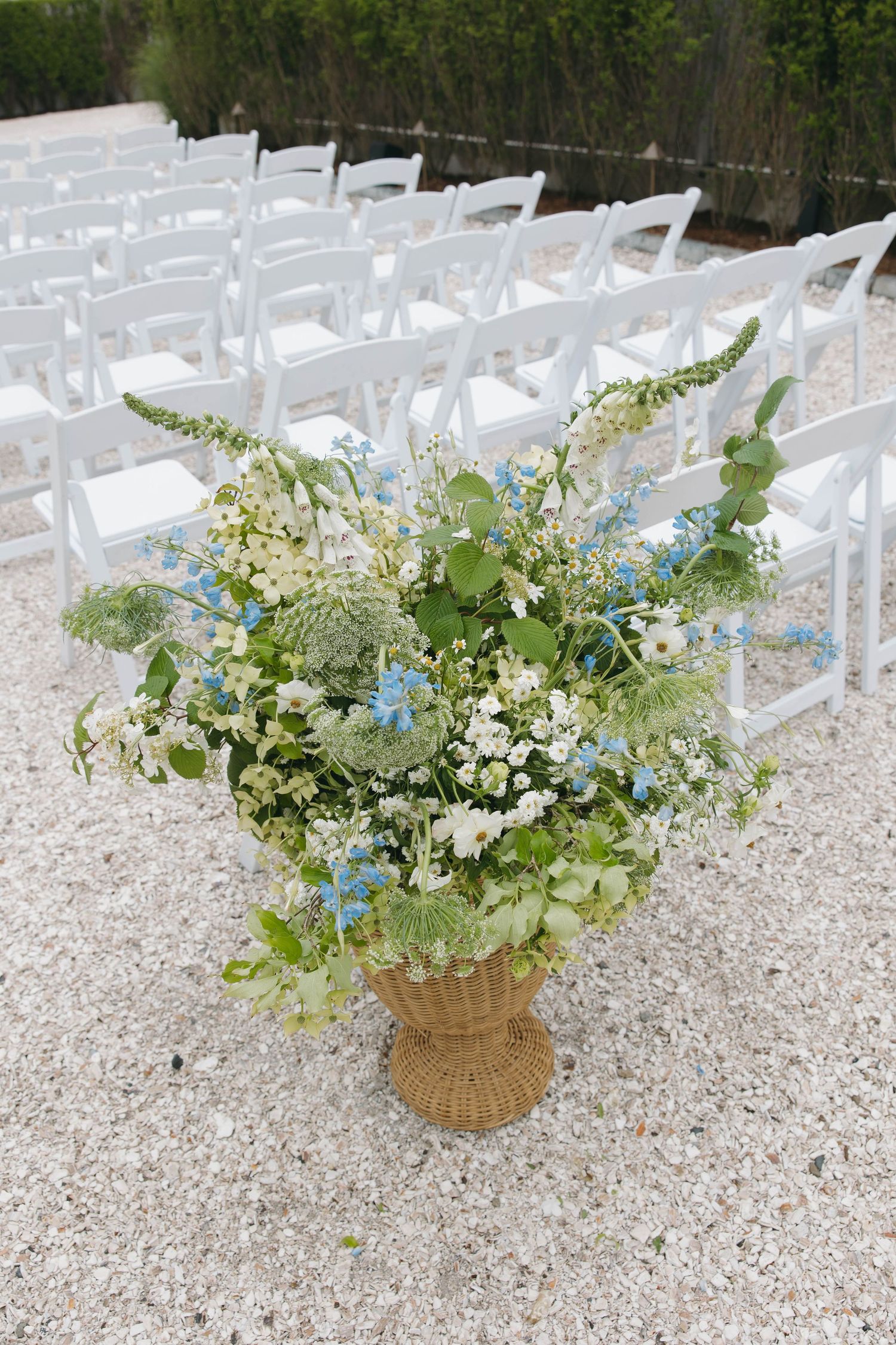 White floral arrangements in woven baskets line the aisle at an outdoor wedding ceremony.