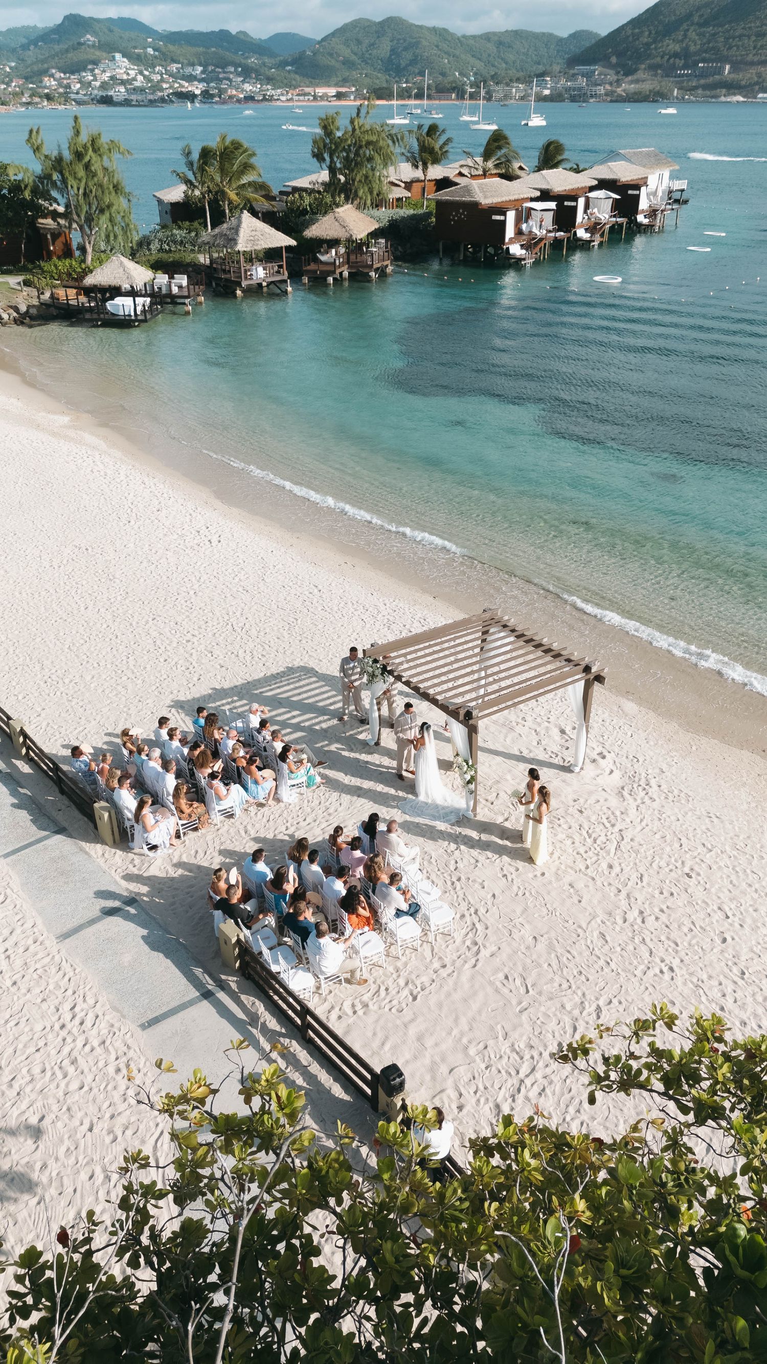Aerial view of an intimate beach wedding ceremony with guests seated on white sand beside turquoise Caribbean waters.