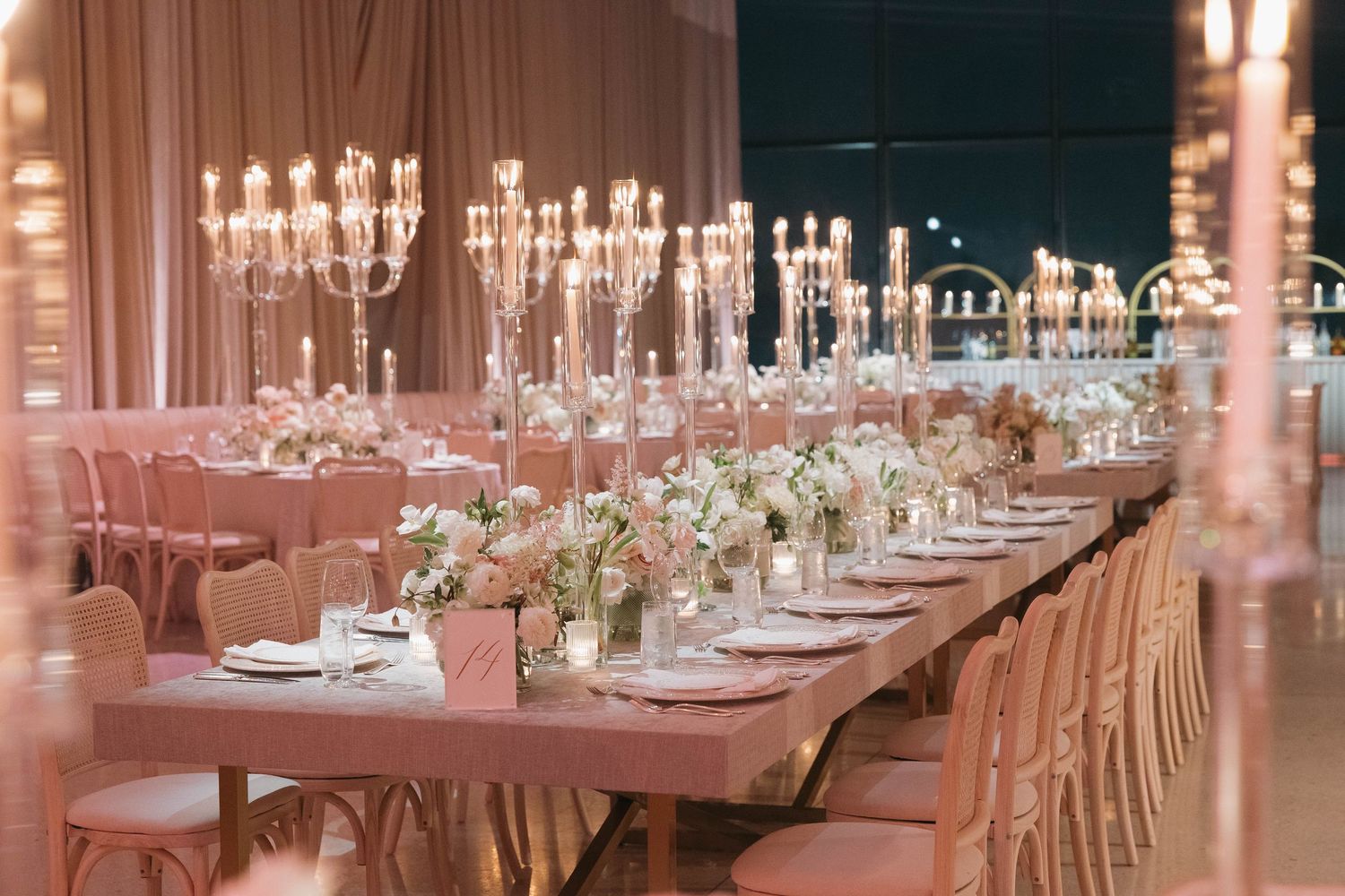 Elegant wedding reception table with crystal chandeliers, pink chairs, and white floral centerpieces in a romantic setting.