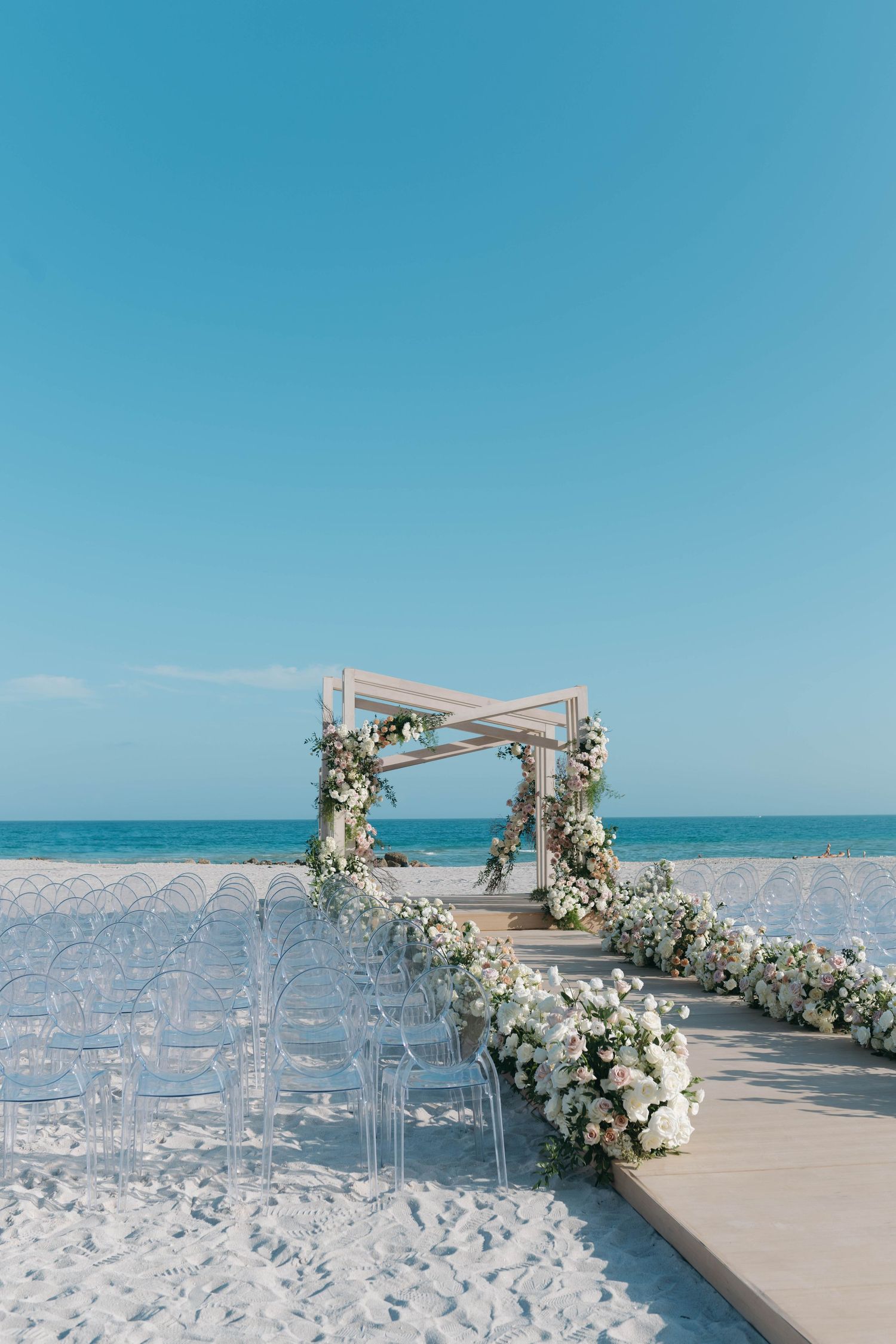 Beach wedding ceremony setup with clear chairs and white floral decorated arch on pristine sand.