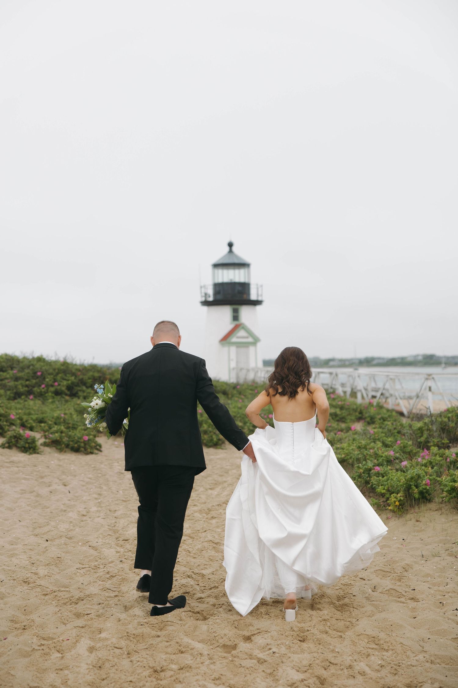 Couple walking on sandy beach path near lighthouse on overcast day.