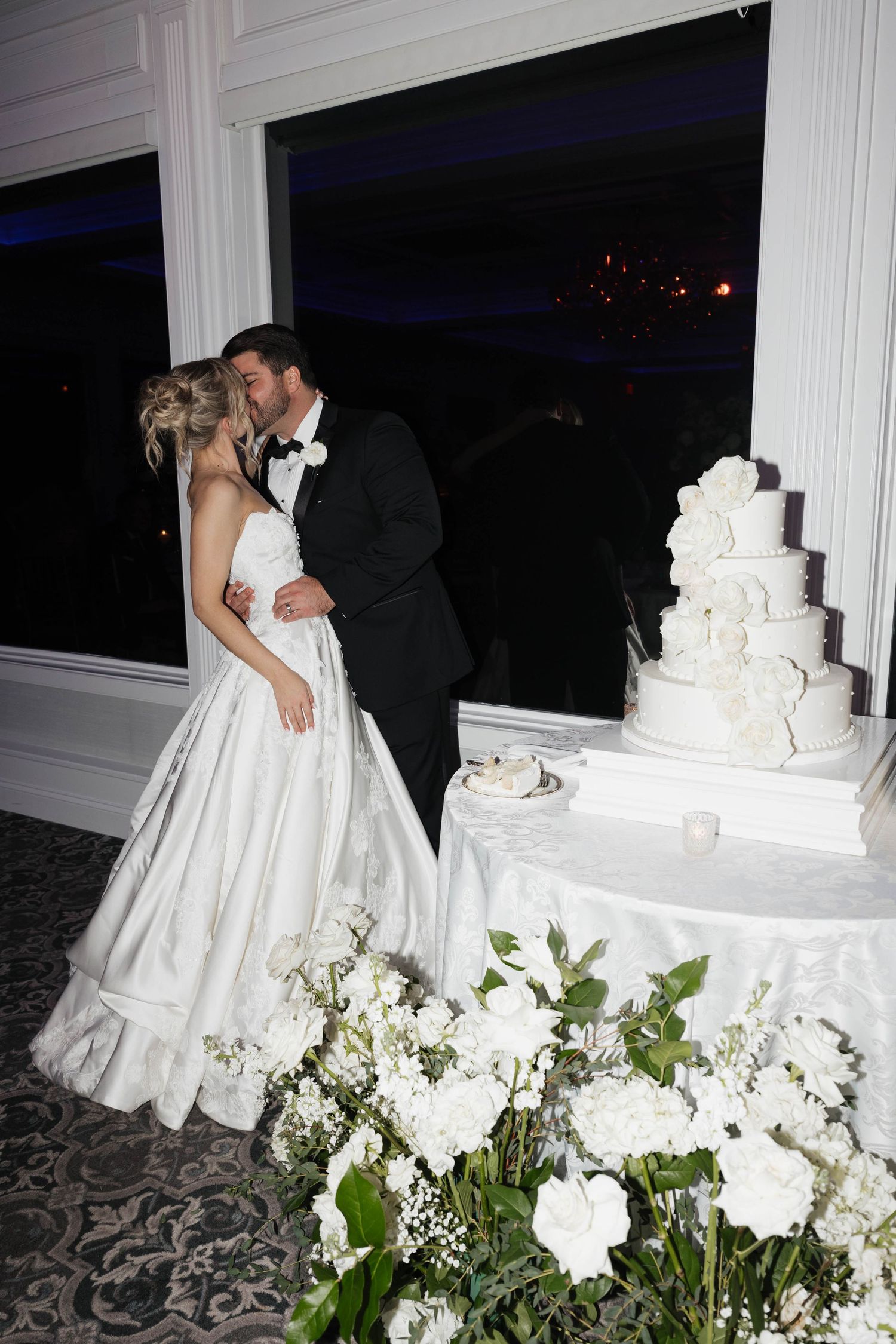 Wedding couple sharing intimate moment by their white tiered cake surrounded by floral arrangements.