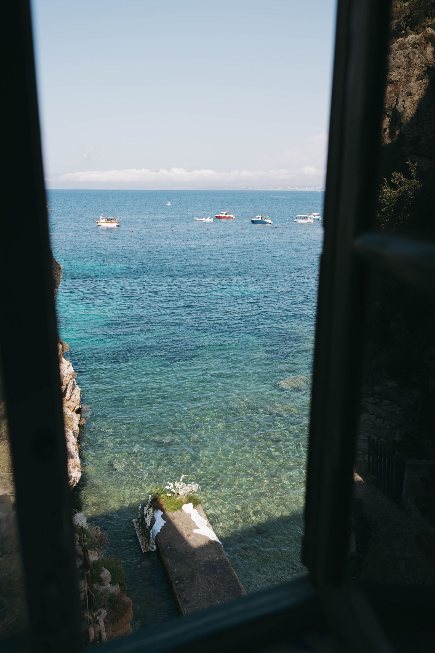 View through a dark window frame of sparkling Mediterranean waters with boats floating in the distance.