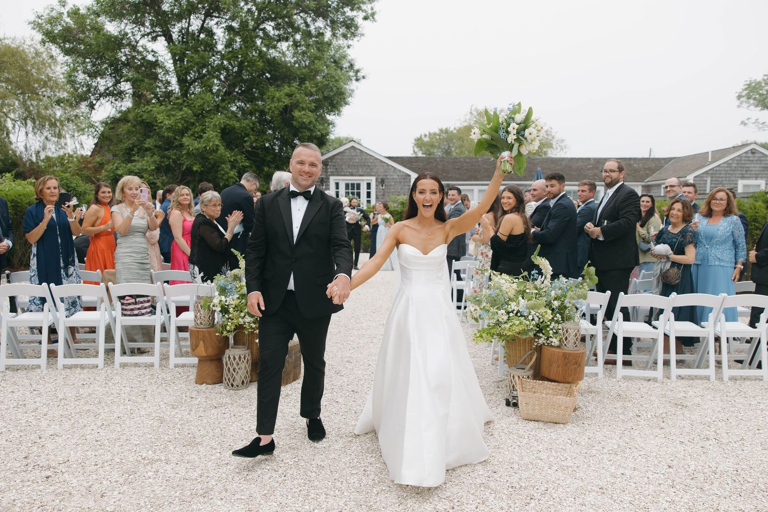 Wedding ceremony guests seated in white chairs on gravel with Cape Cod style houses in background.
