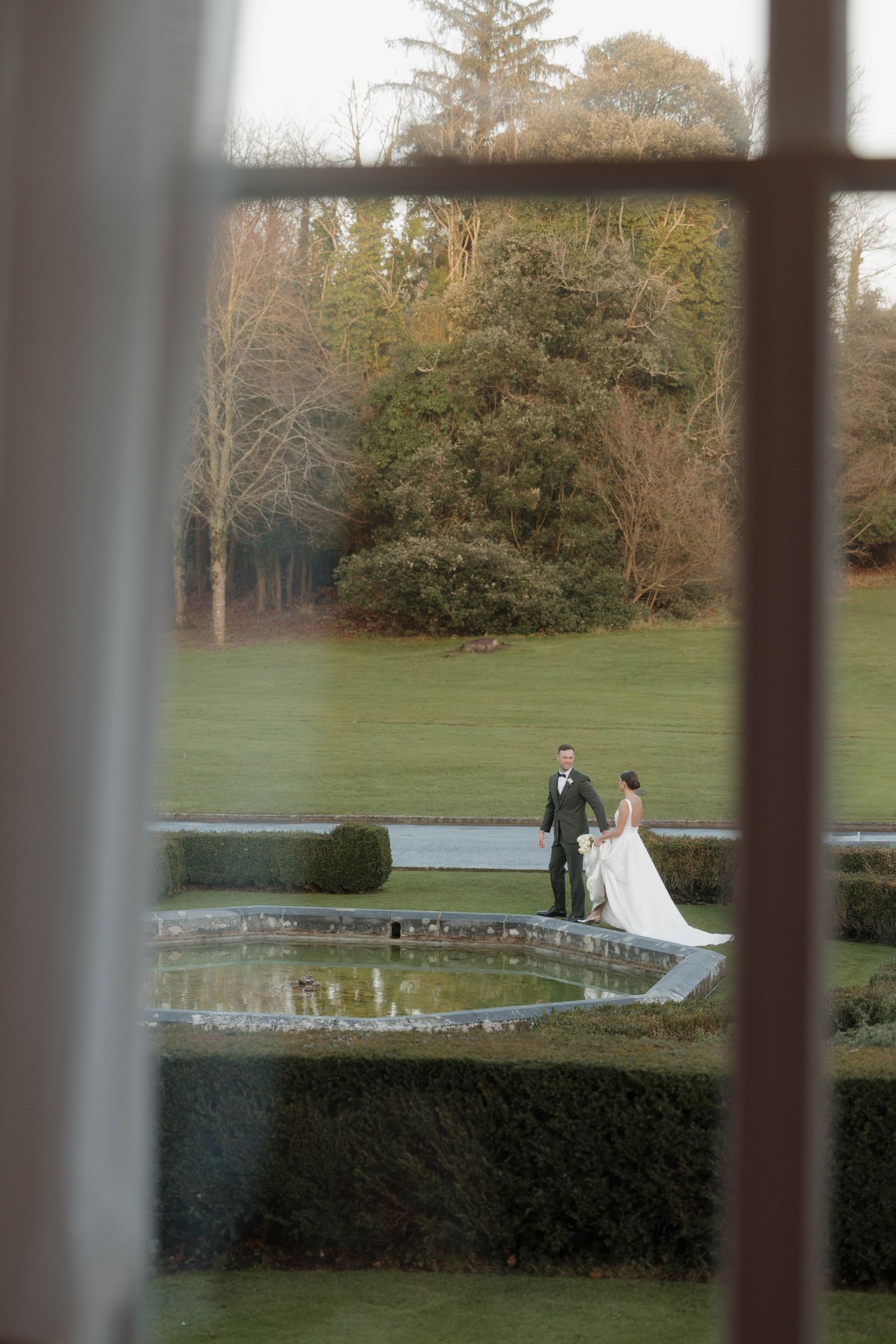 A distant view through windows of a couple in wedding attire by a garden pond.