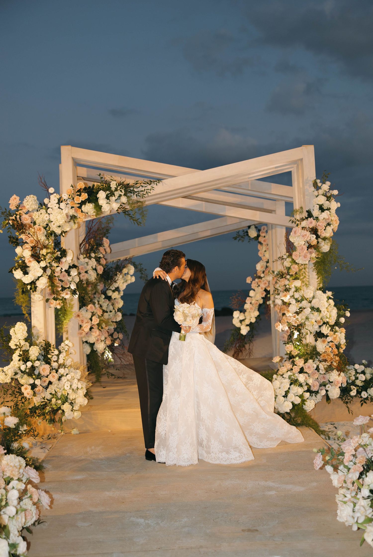 Beach wedding ceremony setup with white floral arch and elegant decorations at dusk.