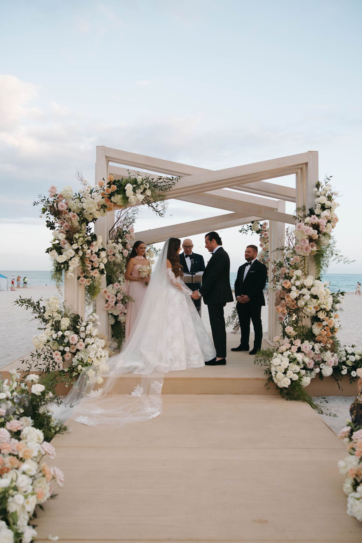 Beachside wedding ceremony setup featuring a modern white pergola decorated with pink and white roses.