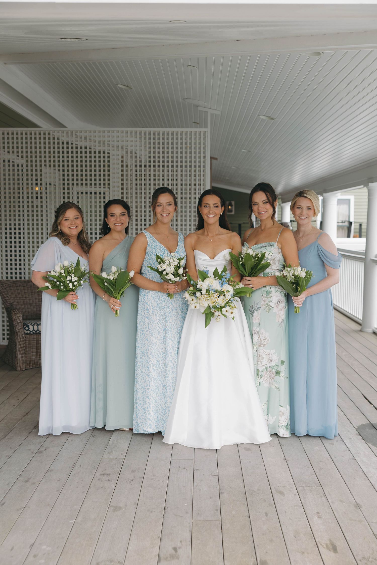 Bridesmaids in light blue dresses pose together on white porch holding floral bouquets.