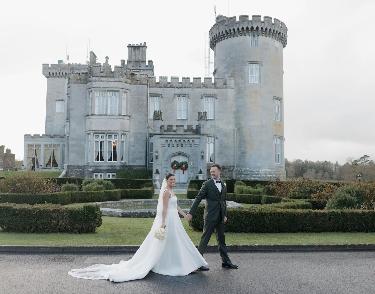 A grand castle with turrets and manicured hedges serves as backdrop for wedding photos.