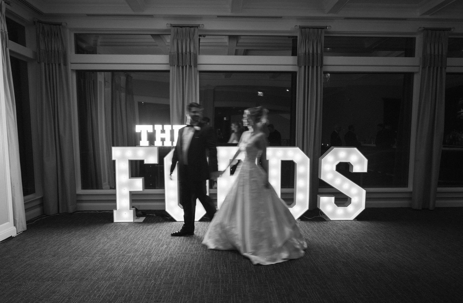 A wedding couple poses in front of illuminated marquee letters spelling 'THE FORDS' at their reception.