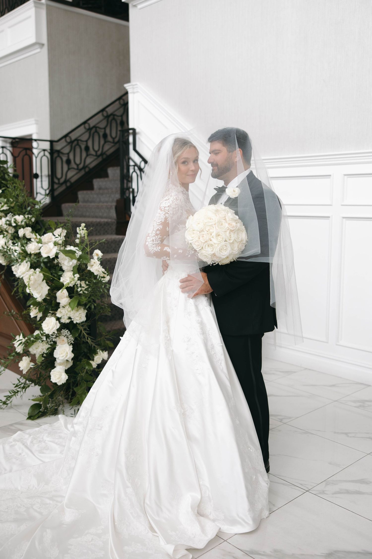 Elegant wedding couple embraces in a white interior space decorated with floral arrangements.