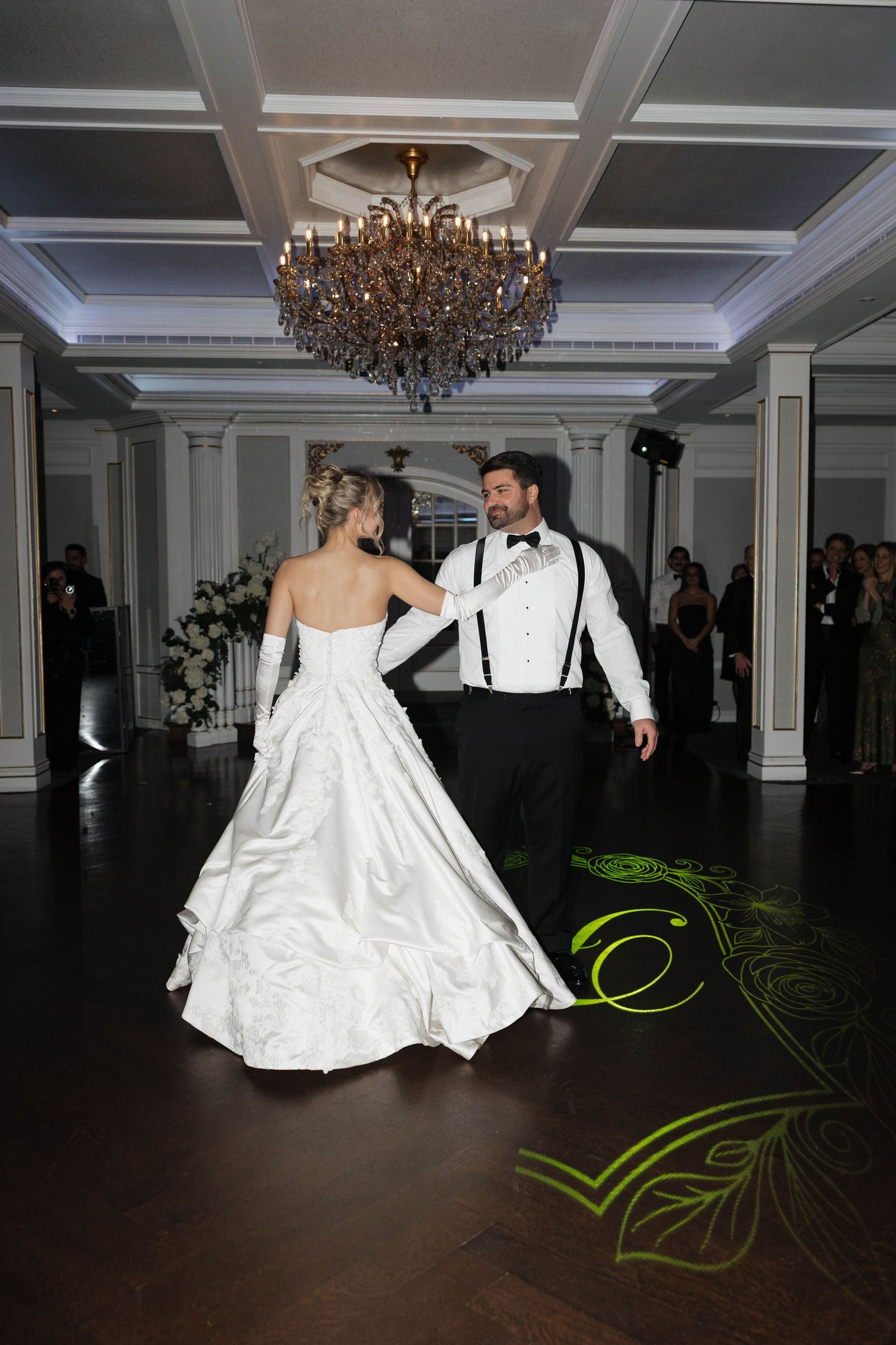 Wedding couple dancing beneath ornate chandelier in elegant ballroom setting.
