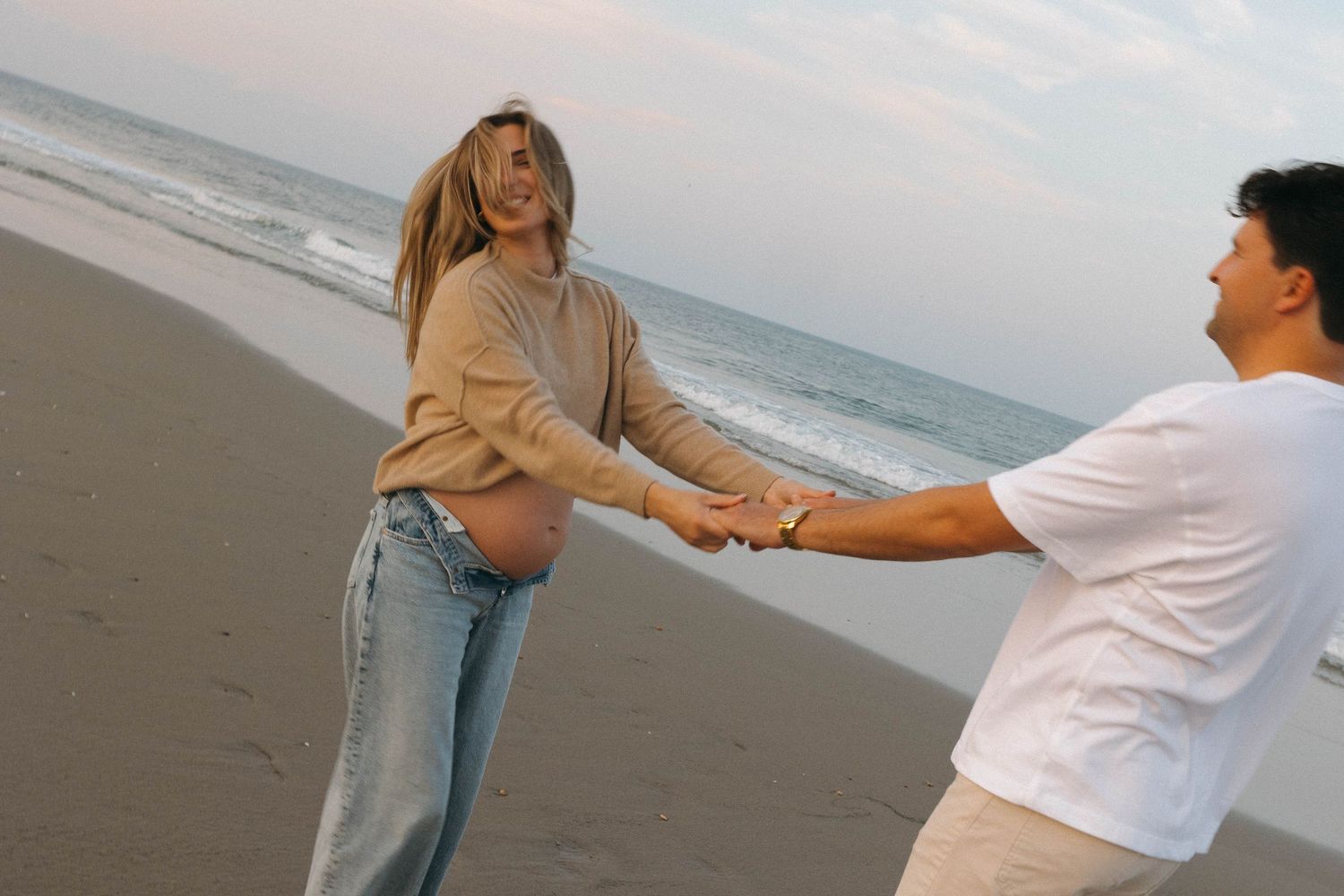 Two people holding hands and playfully interacting on a sandy beach with ocean waves in the background.