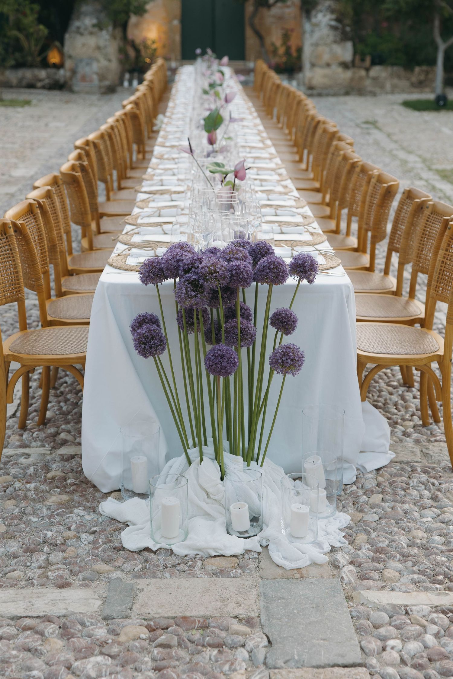 Purple allium flowers decorate a white-clothed dining table with wooden chairs in an outdoor setting.