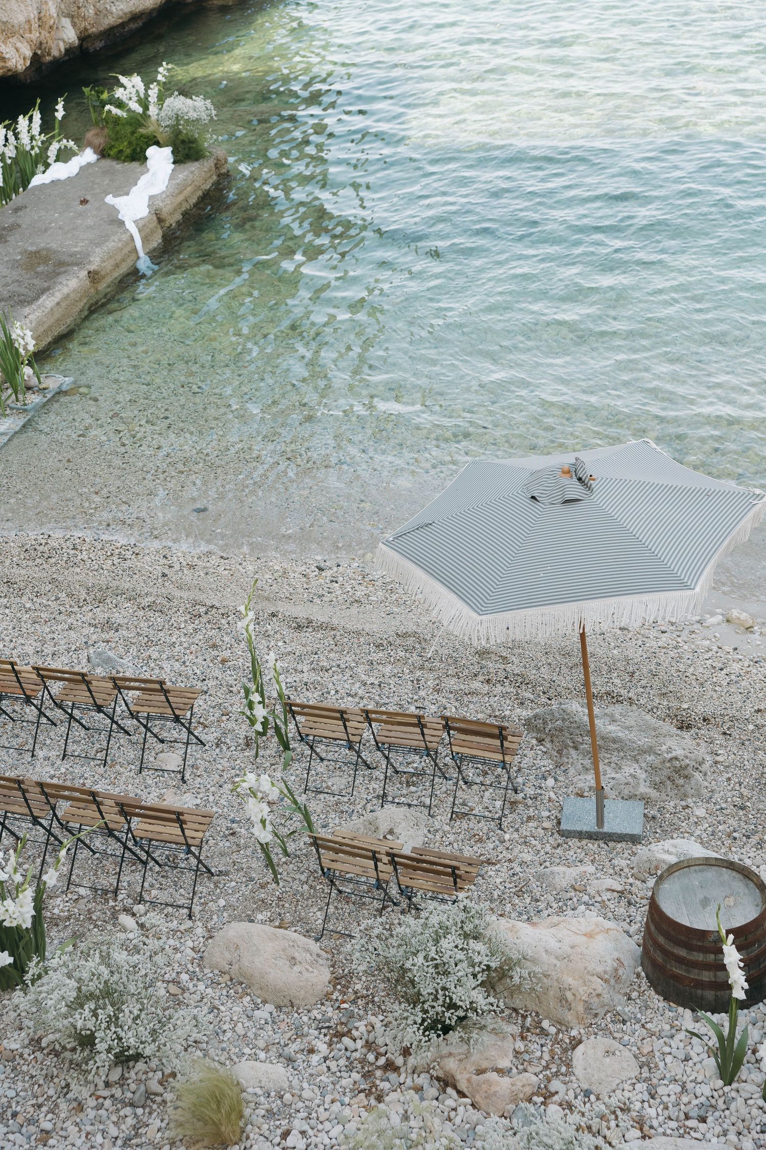 Minimalist beach wedding setup with wooden chairs and grey umbrellas along the shoreline.