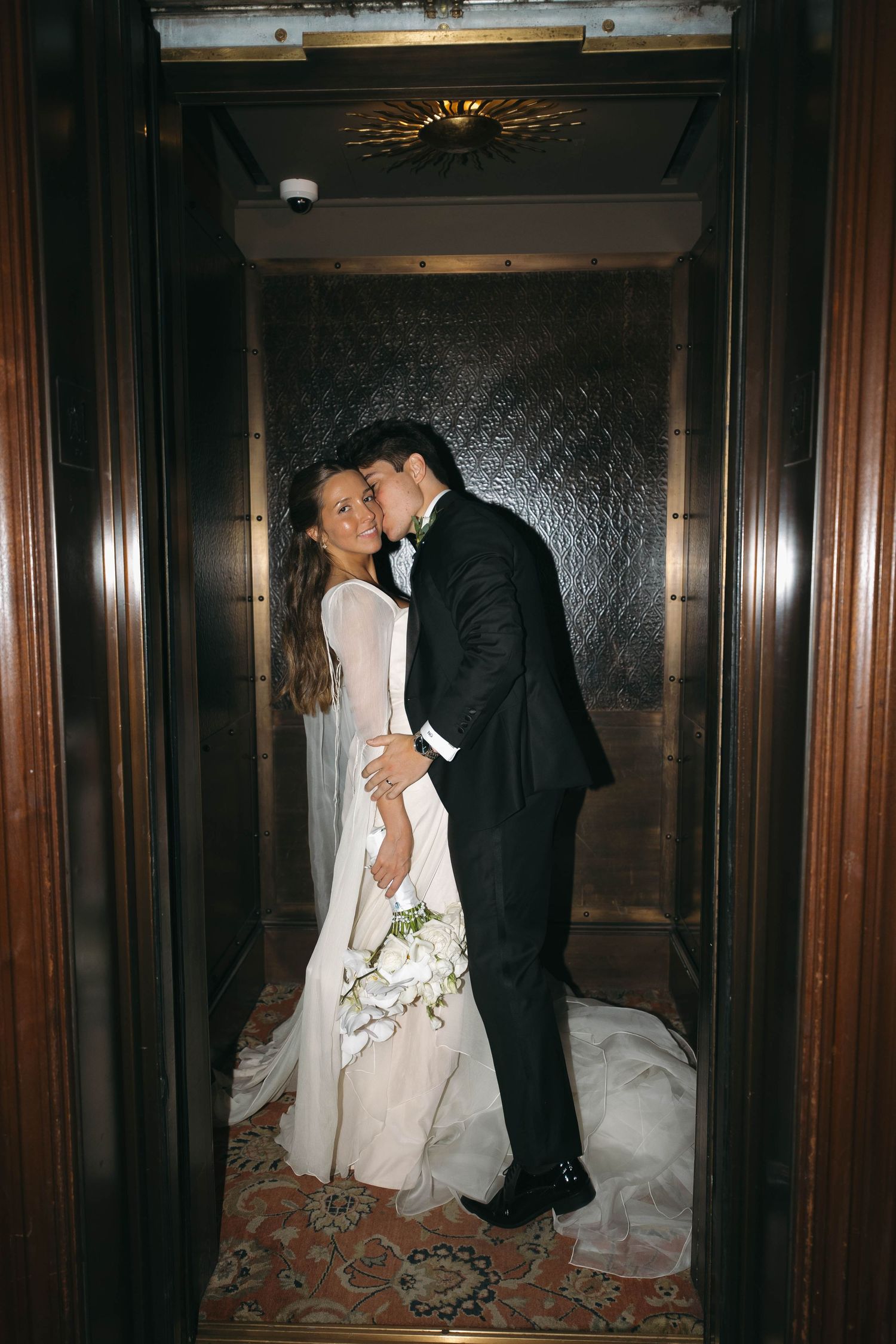 A bride and groom share a romantic kiss inside an elegant elevator, she holds a white floral bouquet in her wedding dress.