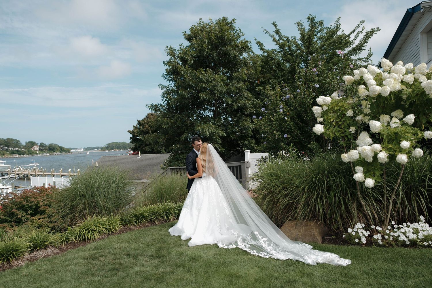Bride and groom share a romantic kiss in a lush garden by the waterfront, with white hydrangeas blooming nearby.