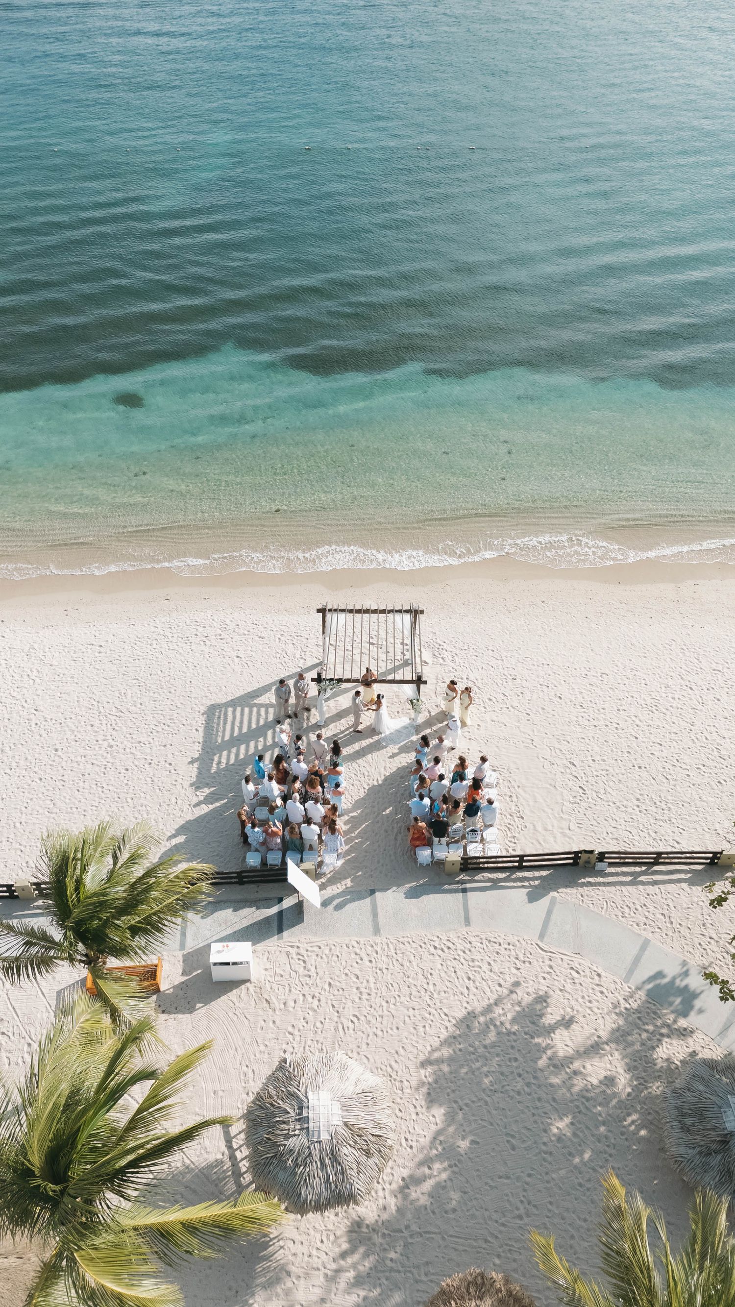 Aerial view of an intimate beach wedding ceremony on white sand with turquoise ocean waves, guests seated facing a wooden arch.