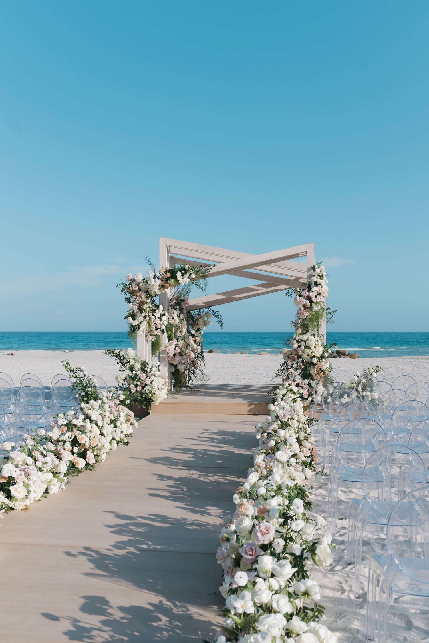 Elegant beachfront wedding aisle with white floral arch and clear ghost chairs.