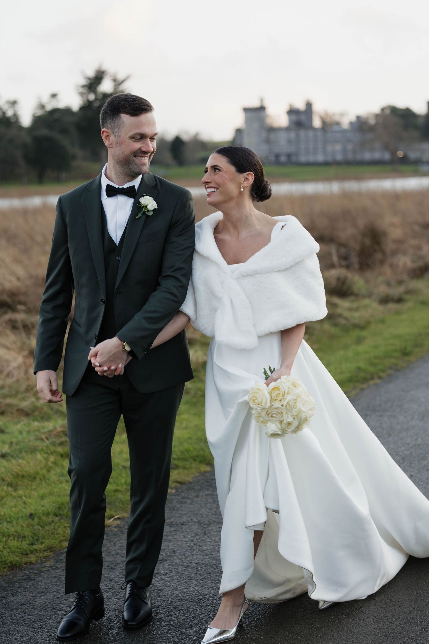 A couple in formal wedding attire shares a joyful moment on a path near a castle.