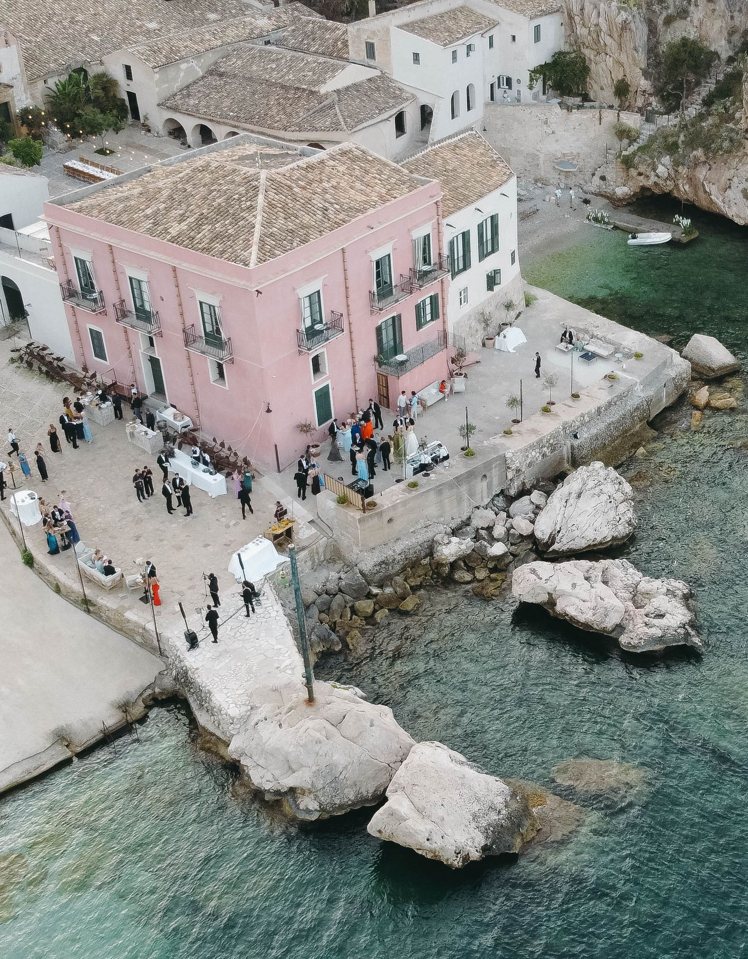 Pink villa perched on rocky coastline with visitors gathering near crystal clear turquoise waters.