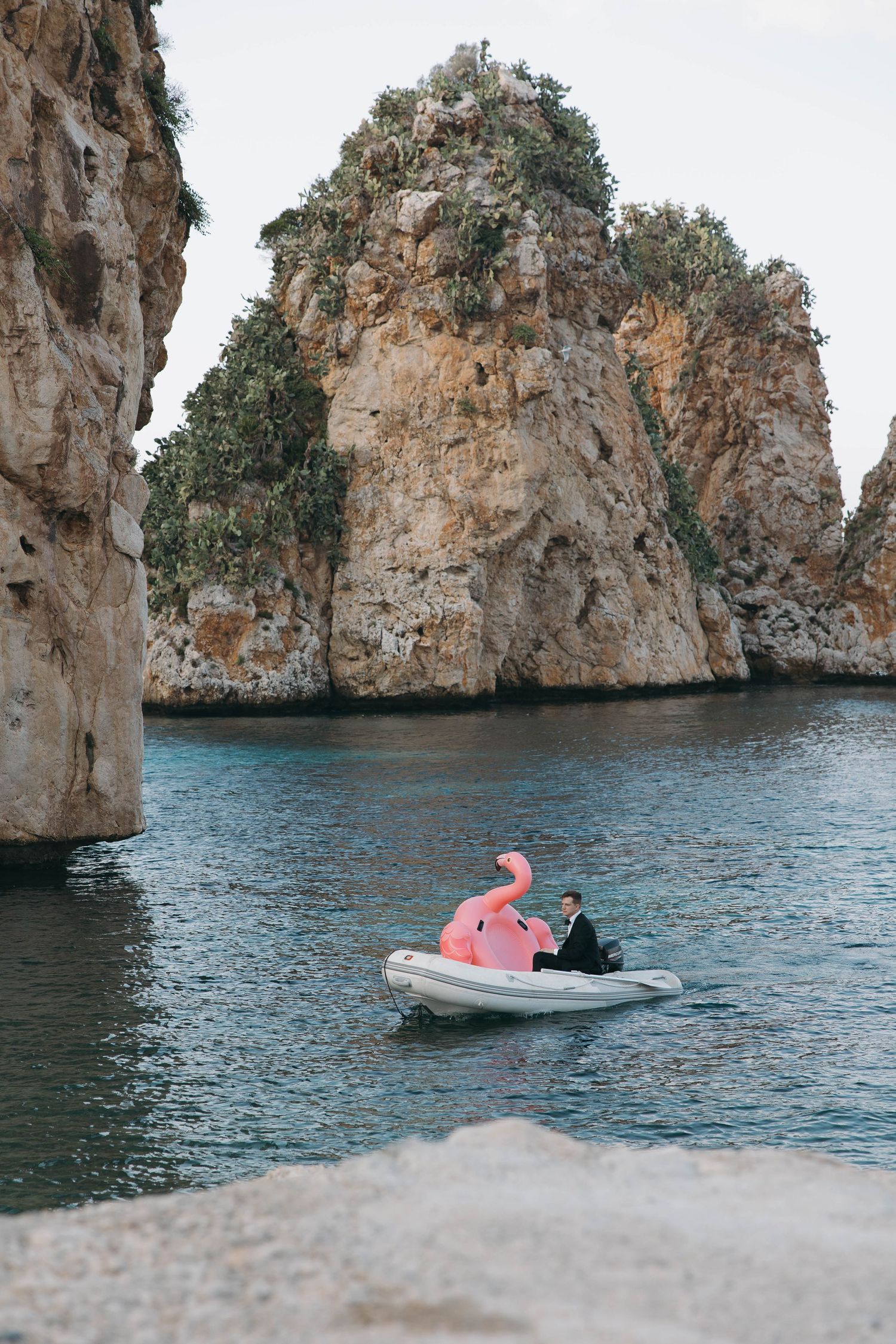 Pink flamingo float drifts near dramatic coastal rock formations in crystal clear waters.