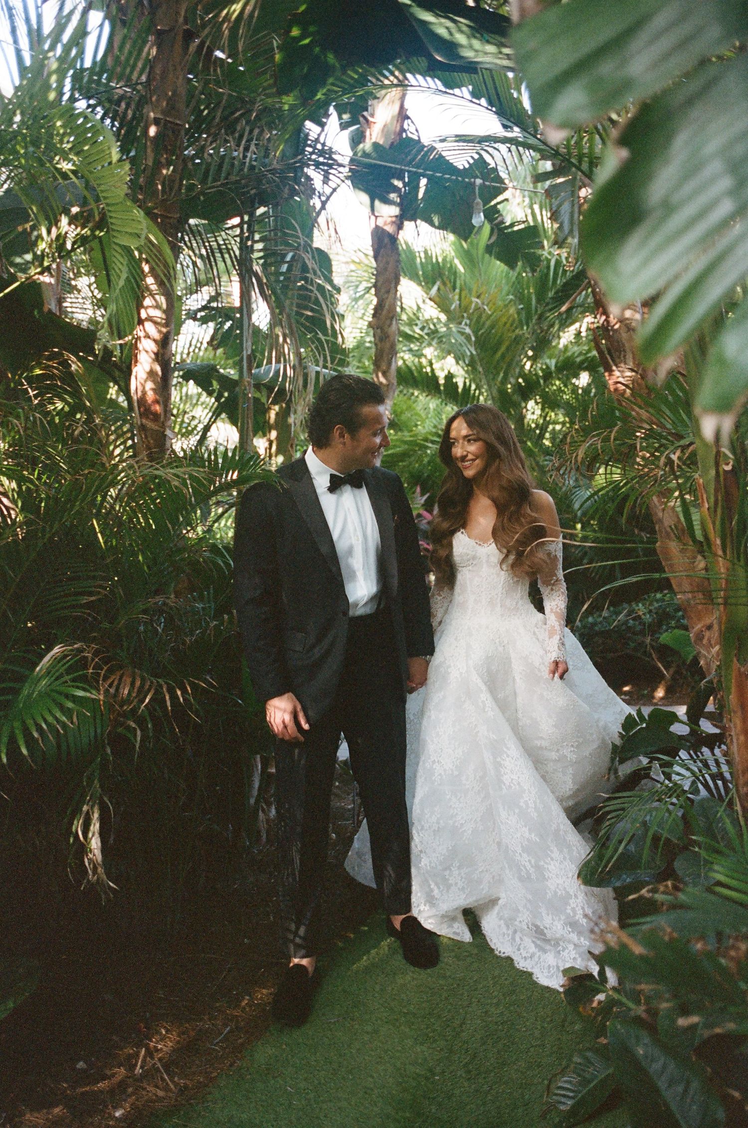 A romantic wedding couple dances together surrounded by lush tropical plants and greenery in a garden setting.