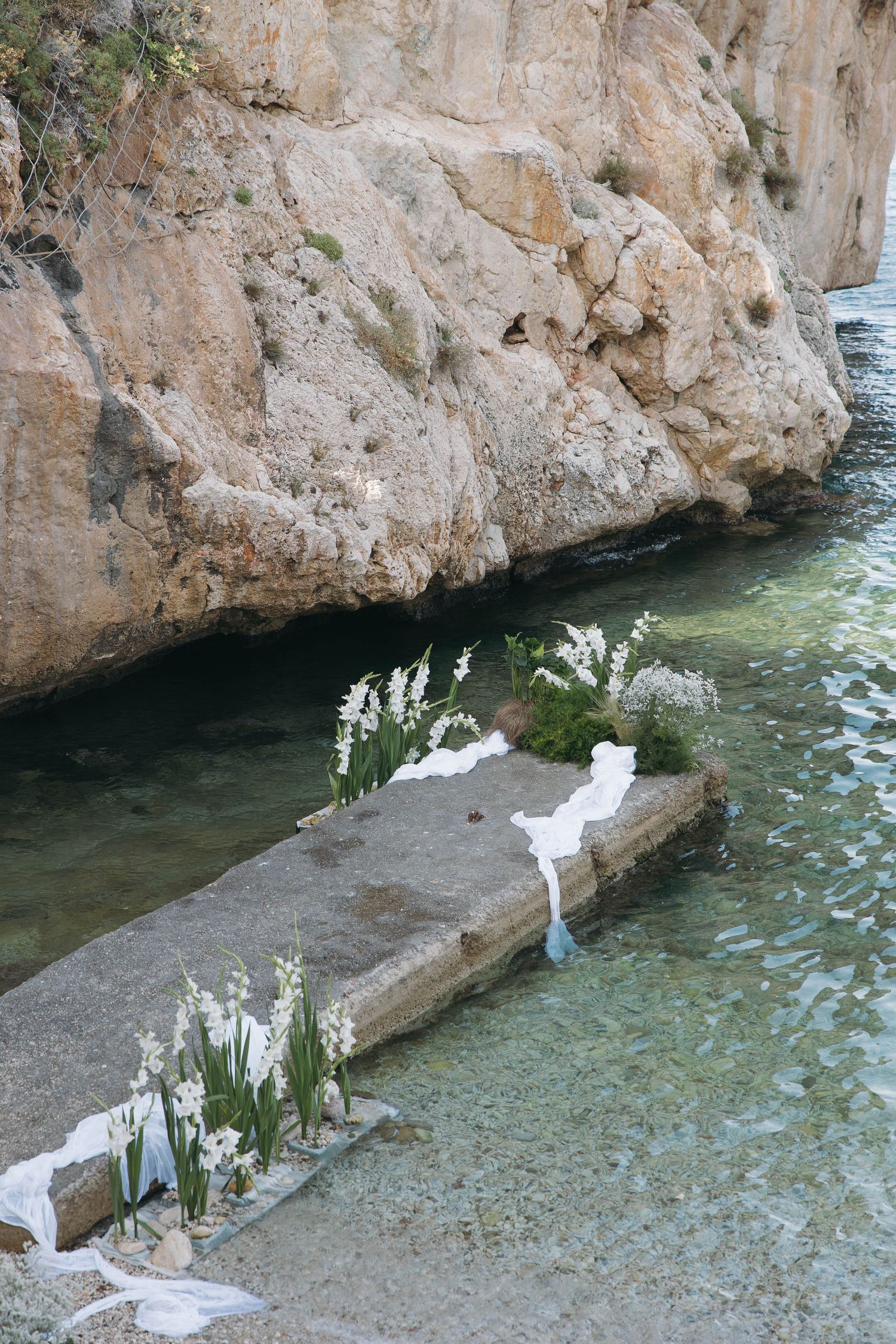 White floral arrangements line a stone pathway along turquoise Mediterranean waters.