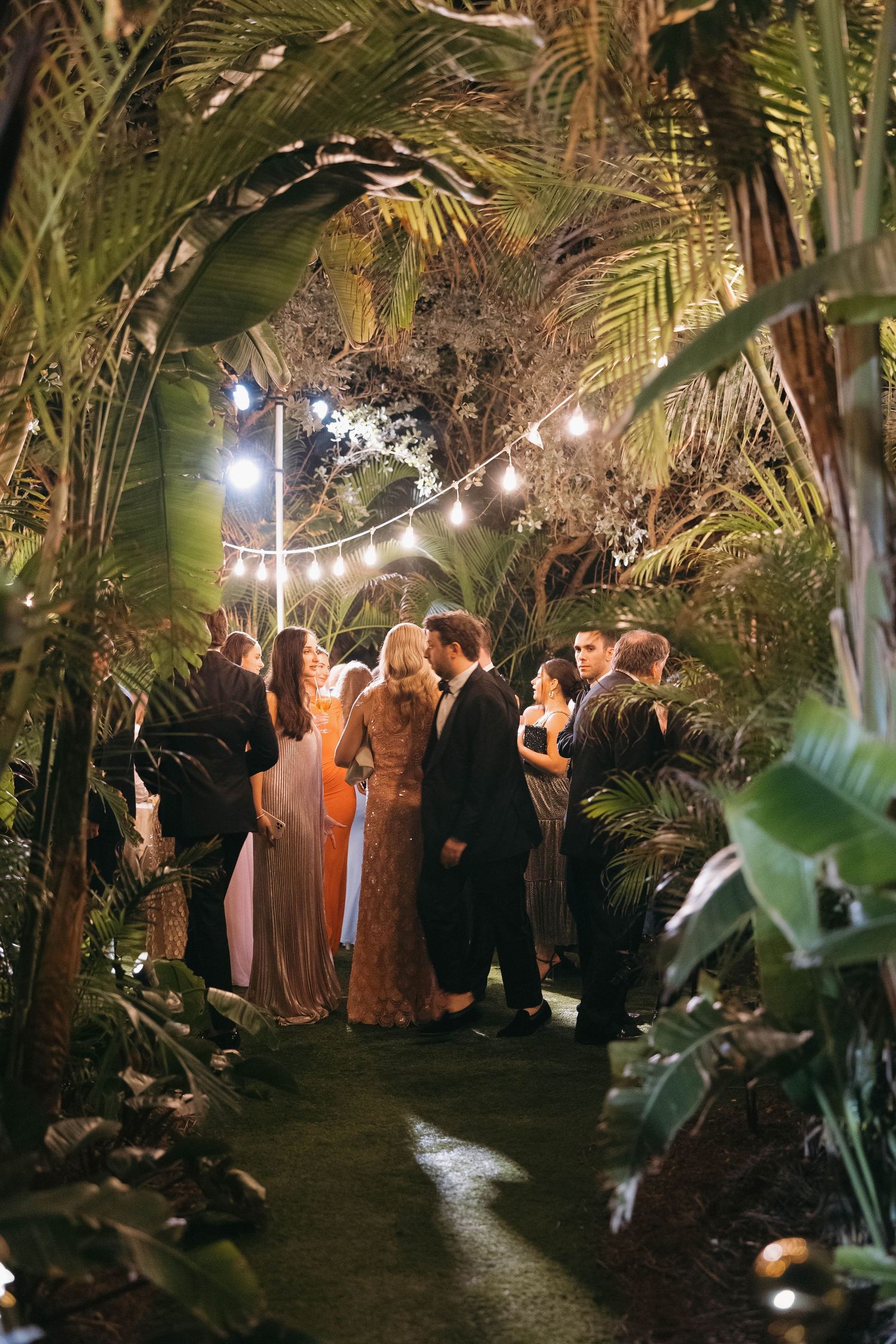 Wedding guests mingle under string lights in a lush tropical garden setting.