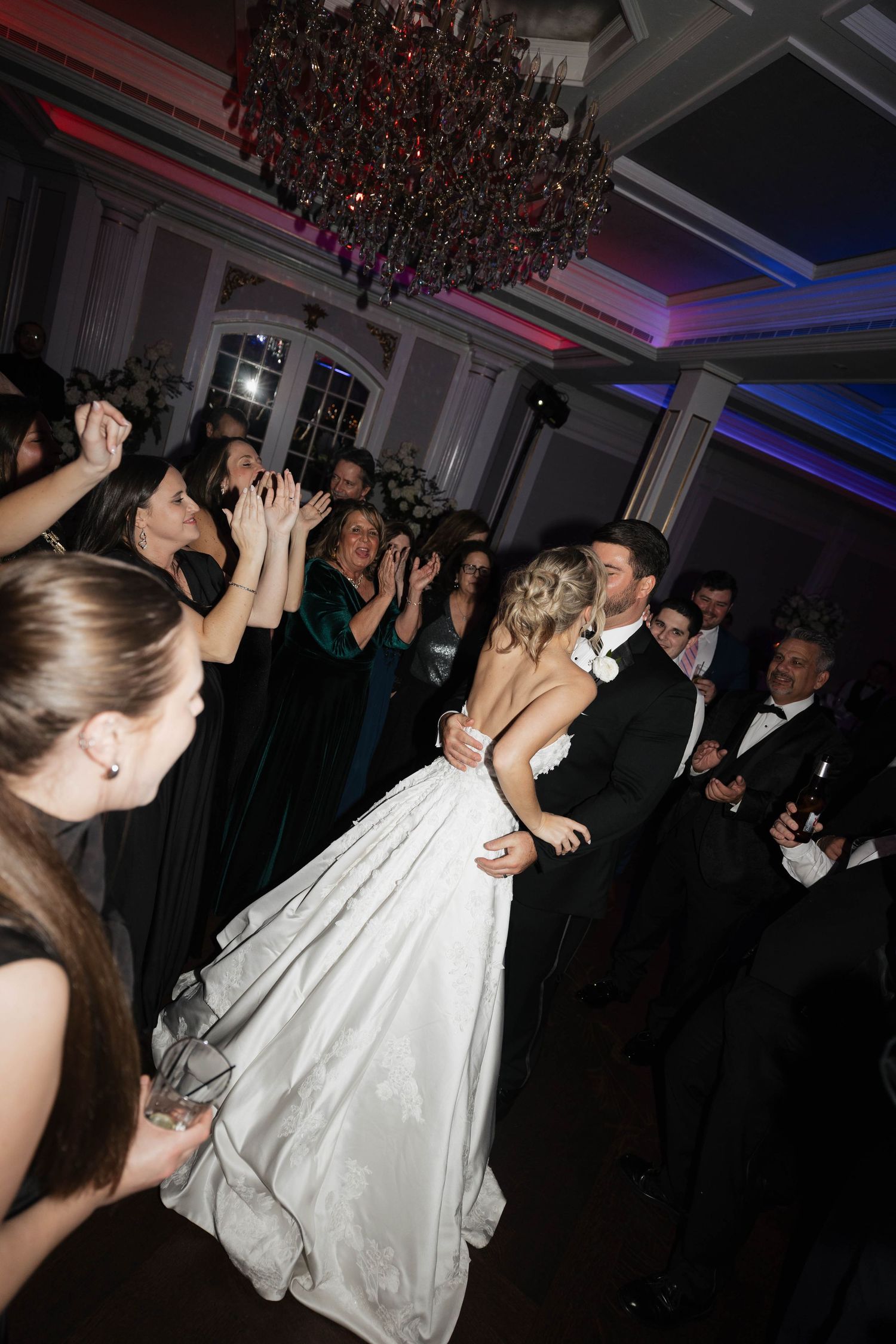 Bride in white gown dances with wedding guests under dramatic chandelier lighting.