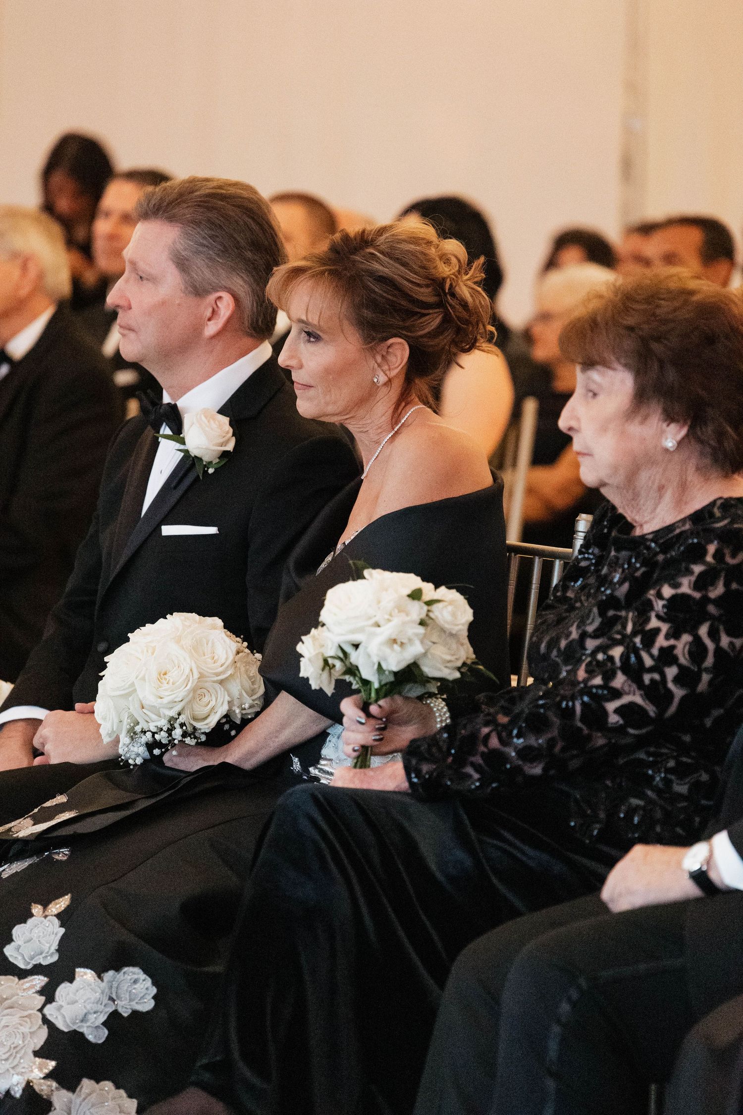 Wedding attendees in formal black clothing holding white bouquets while seated.