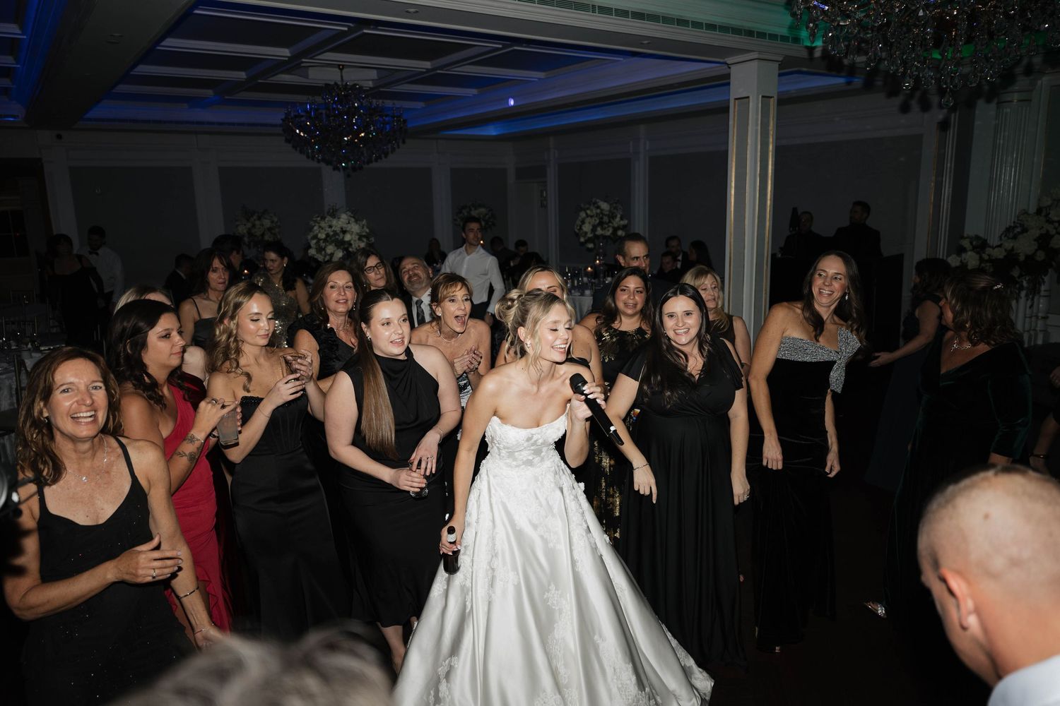 Wedding party dancing and celebrating in a dimly lit ballroom with decorative lighting.
