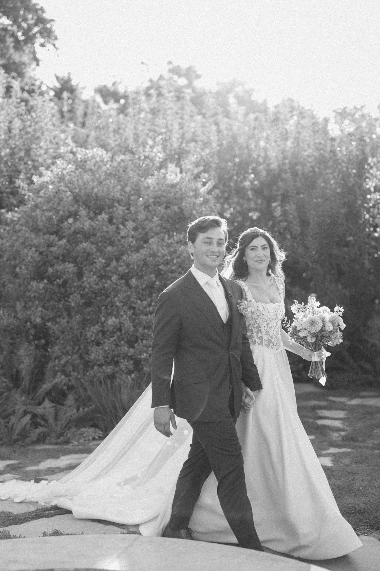 Newlyweds walk together through a garden setting with the bride's white dress train flowing behind them.