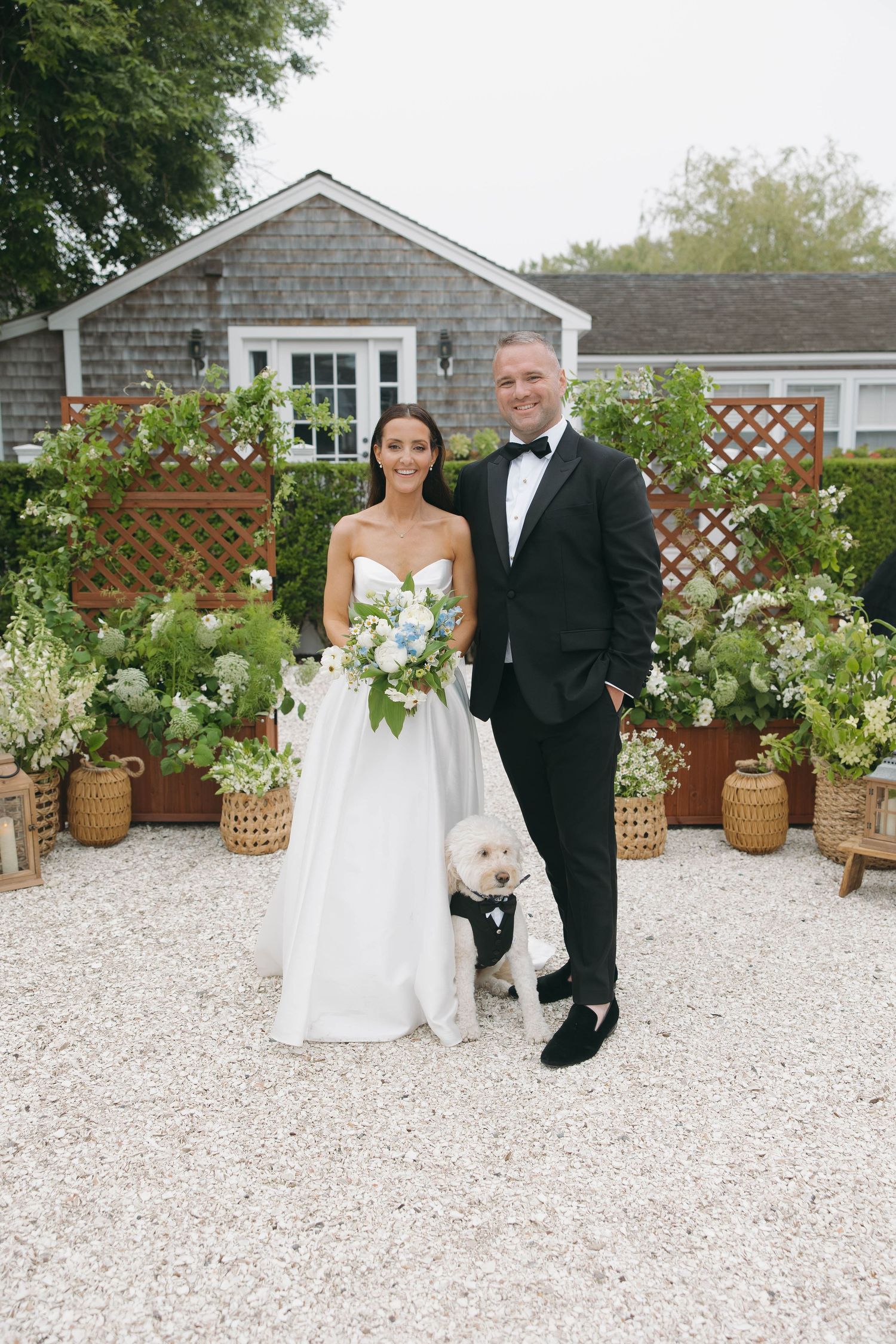 Elegant wedding couple poses in front of a rustic shingle house with potted plants and flowers.