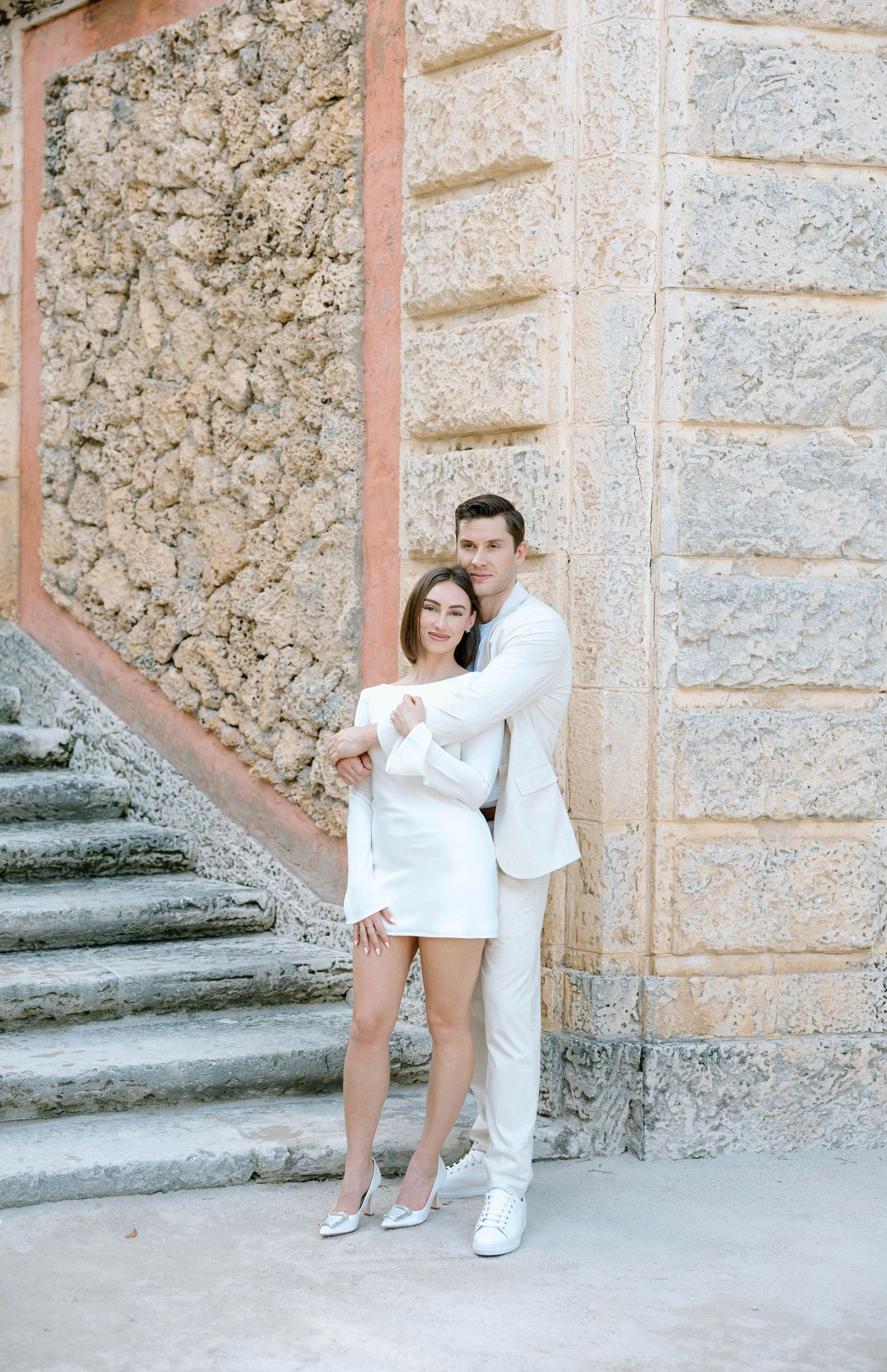 A couple in white outfits poses playfully against a textured stone wall.