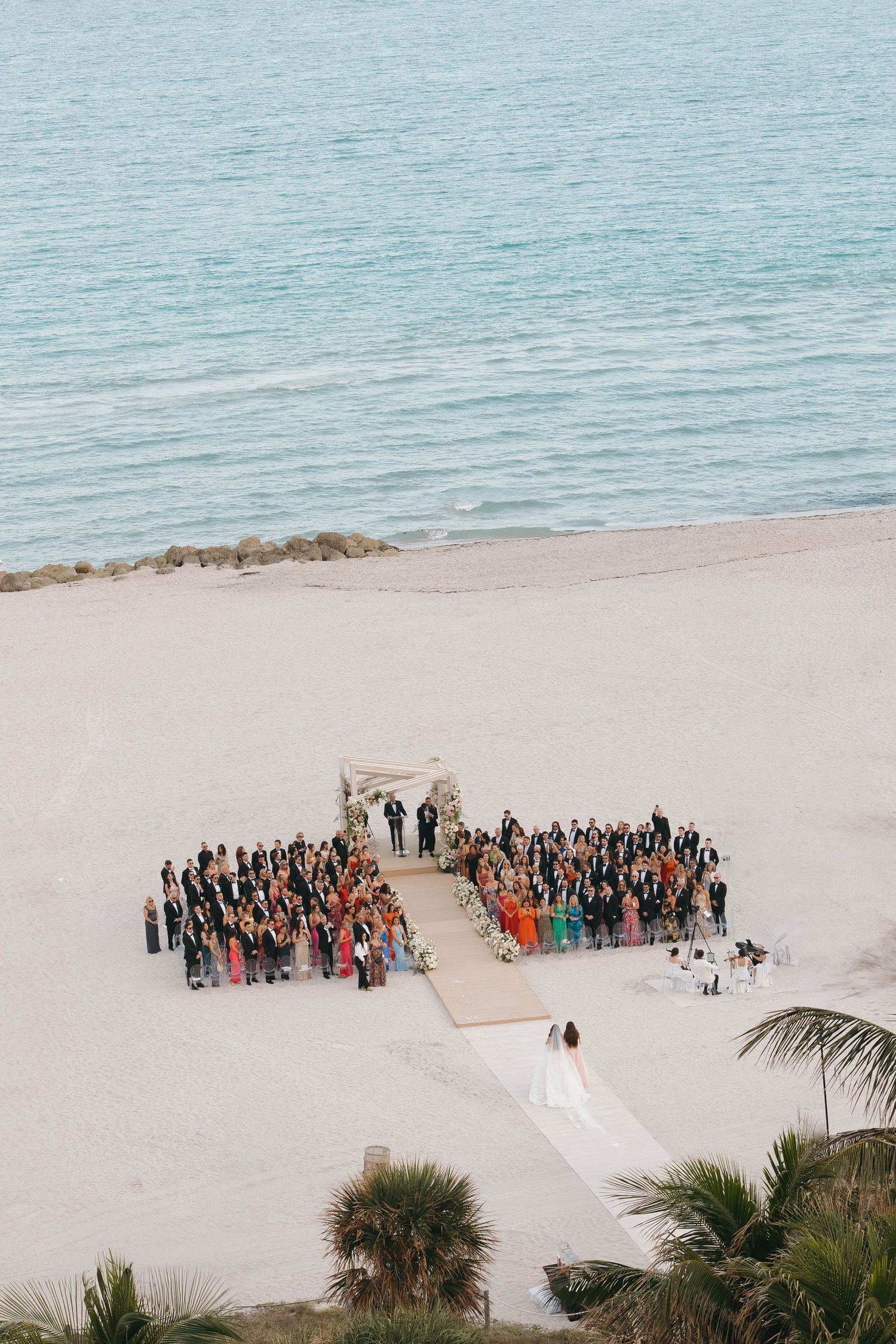 A beach wedding ceremony setup with guests gathered on white sand near turquoise waters.