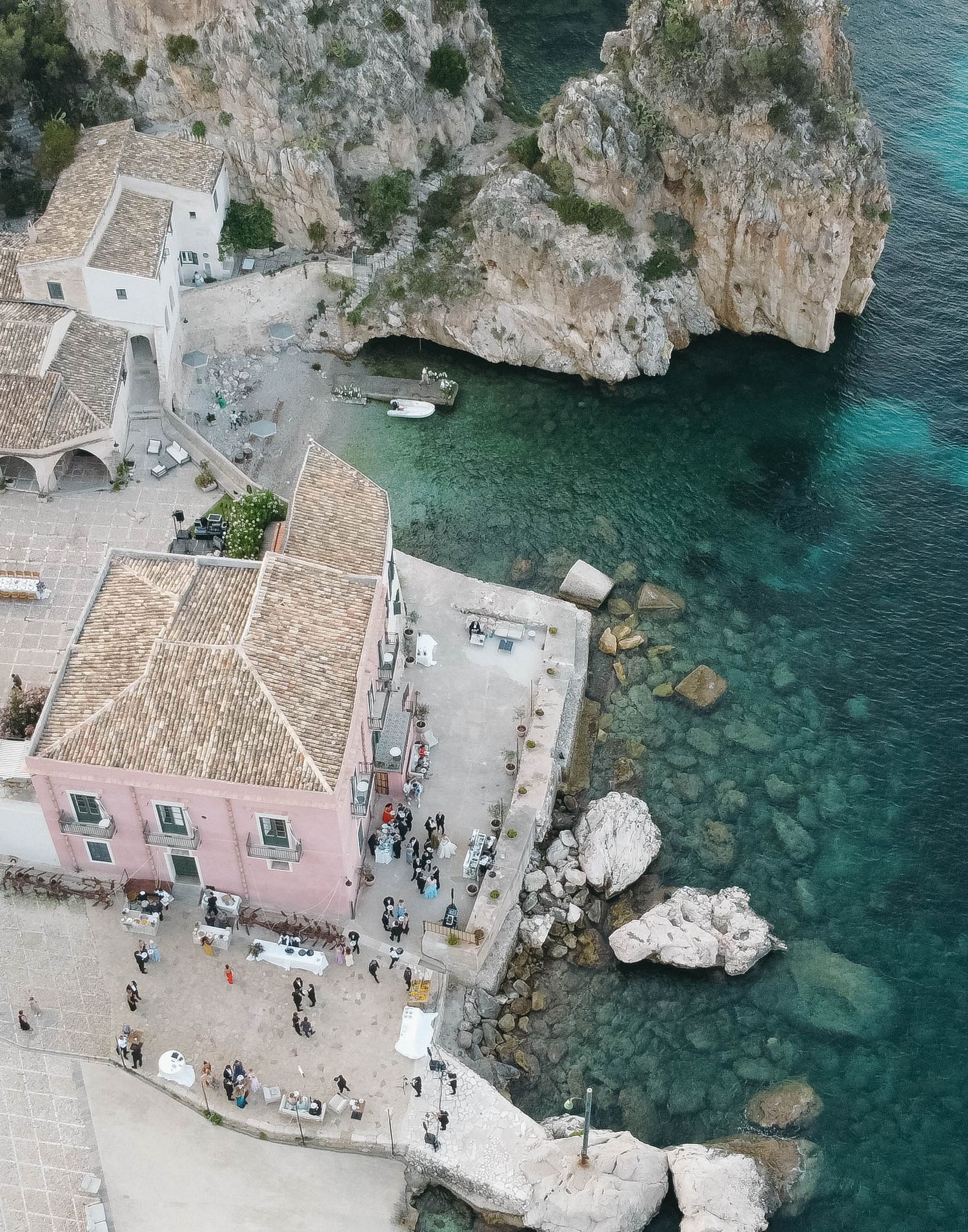 Aerial shot of Mediterranean seaside buildings and gathering crowds along rocky turquoise bay.
