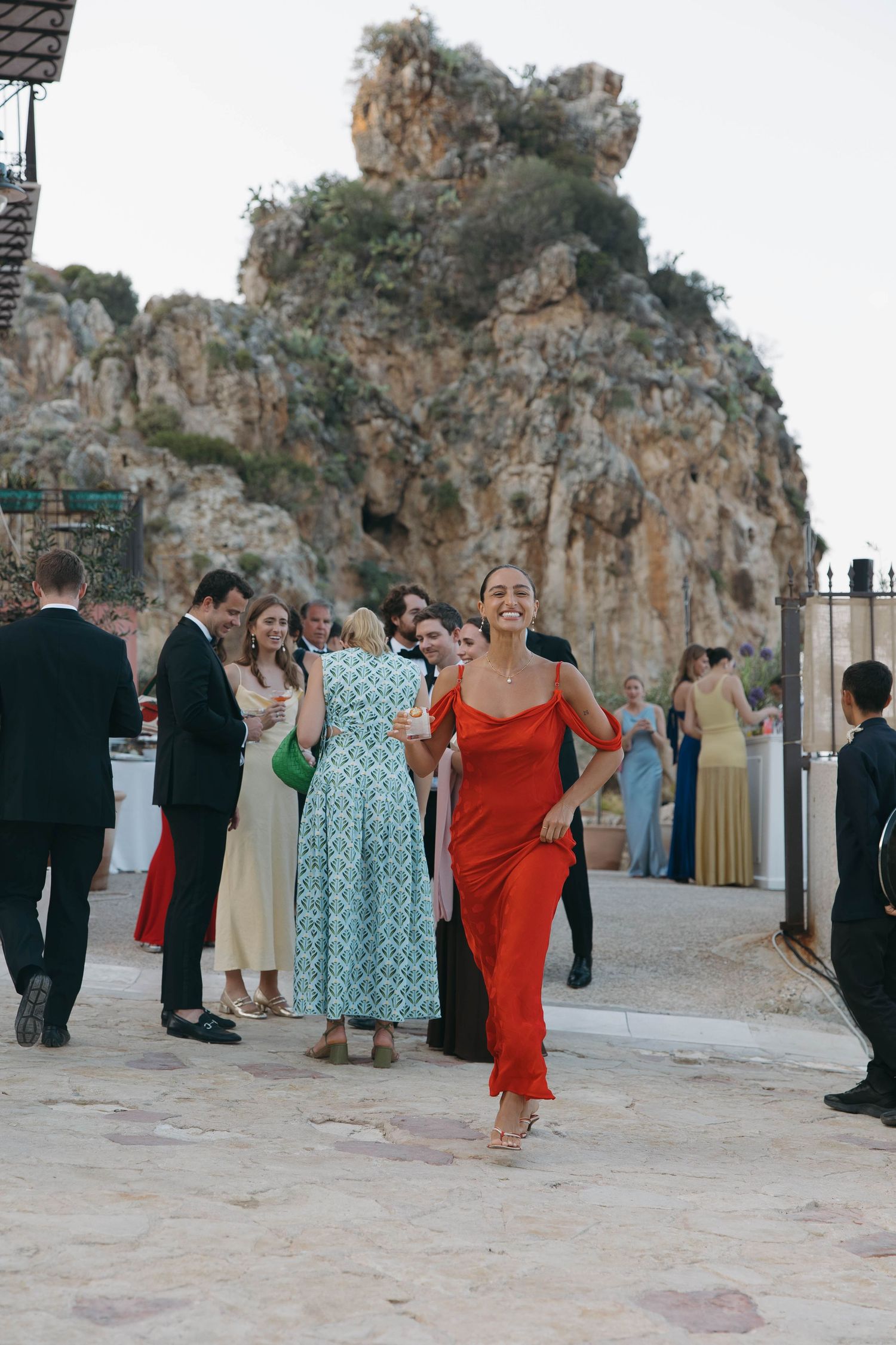 Formal event attendees mingle on stone patio near dramatic coastal cliffs.