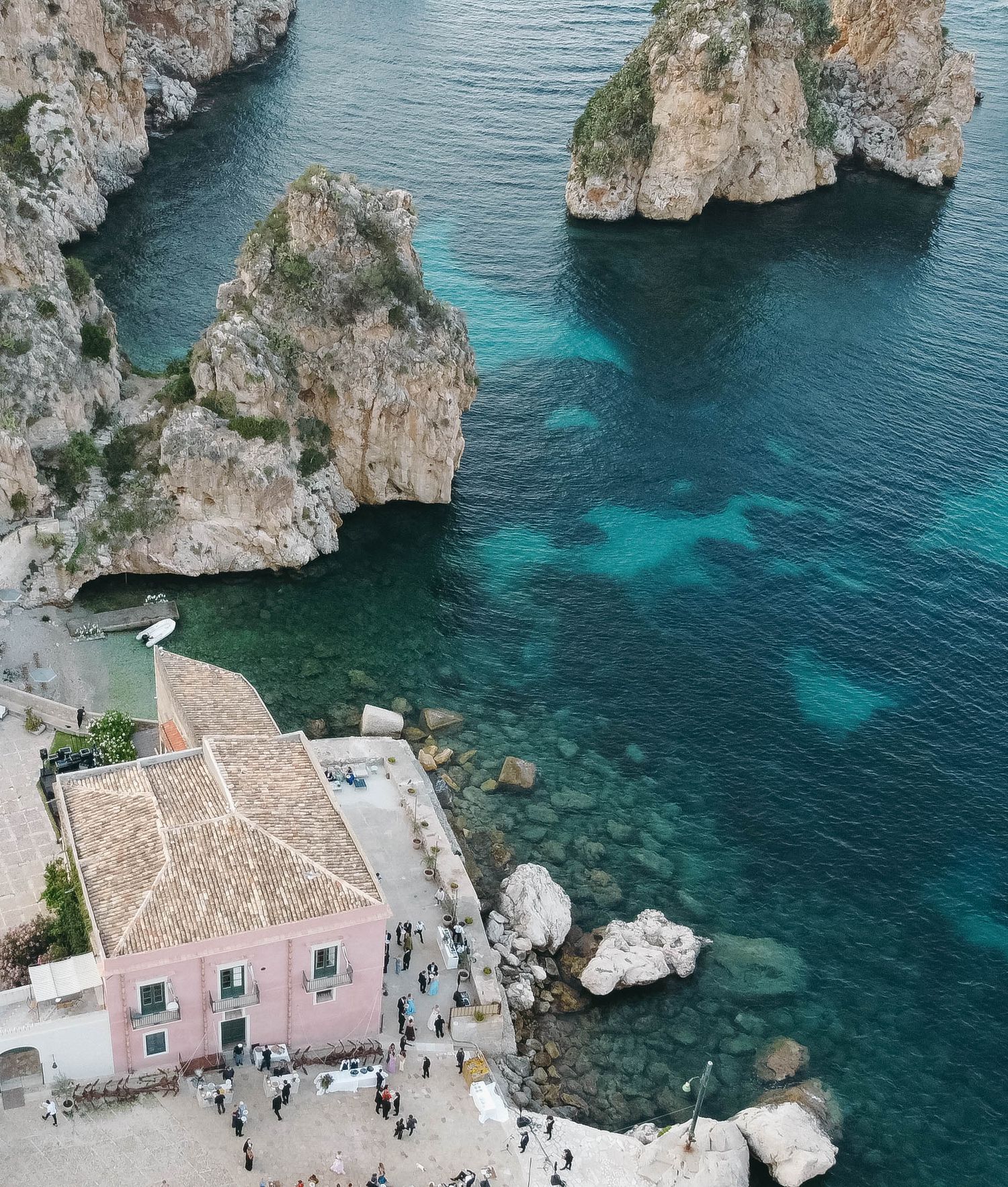 Aerial view of rocky coastal cliffs with turquoise Mediterranean waters and pink buildings near the shore.