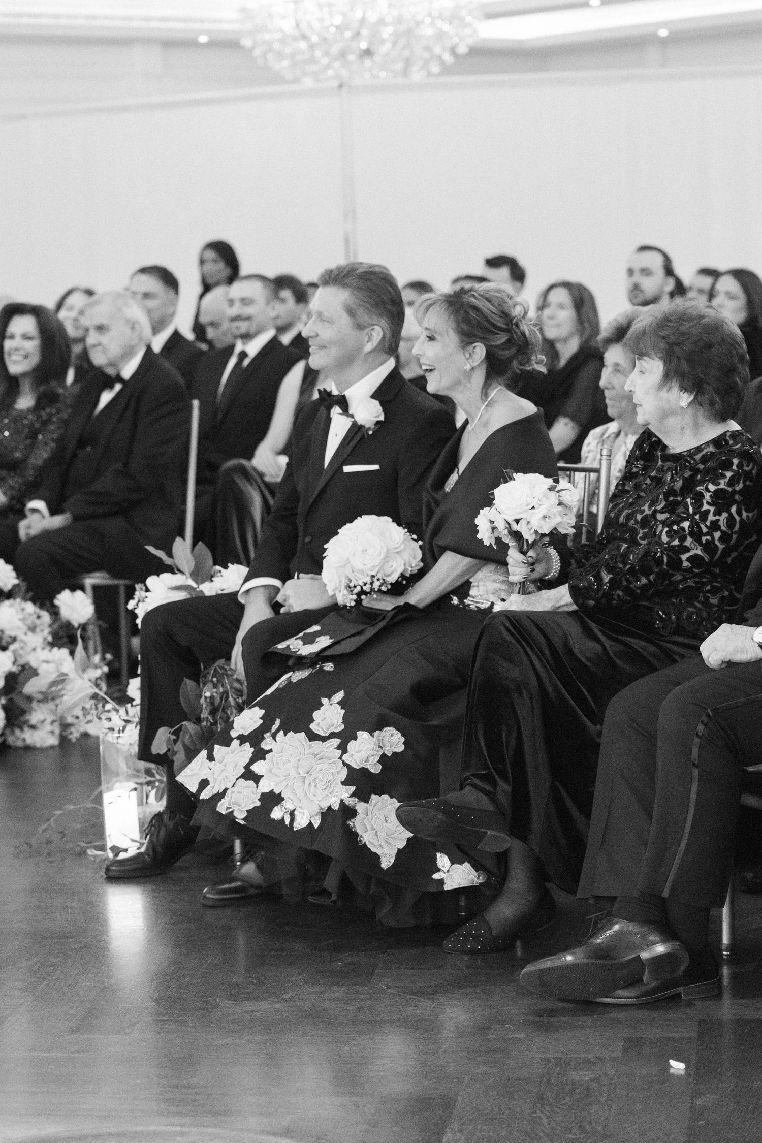 Wedding guests in formal black attire seated in rows with white floral bouquets during ceremony.