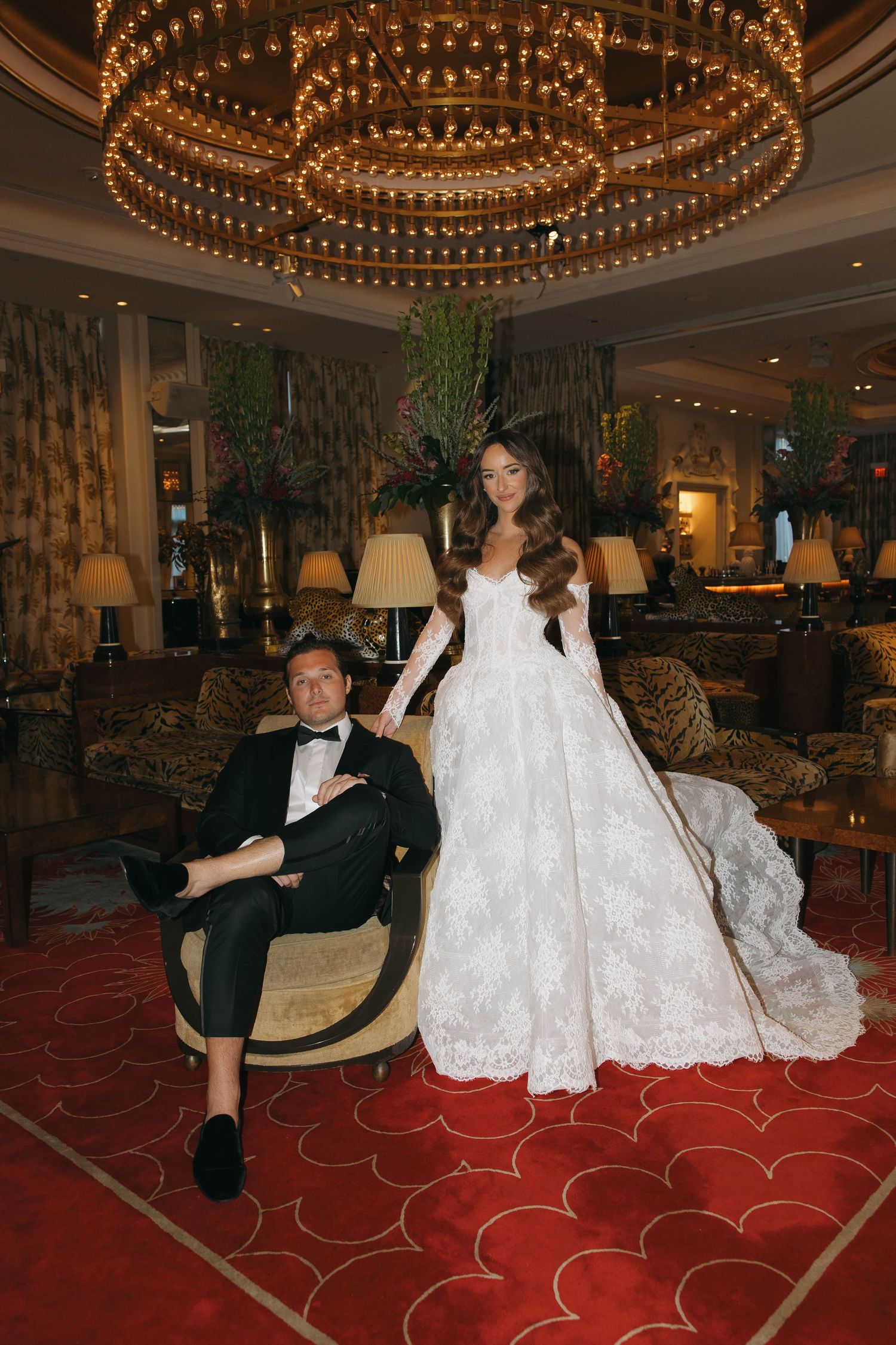 A couple poses playfully under an ornate chandelier on red carpeting in a luxurious venue.