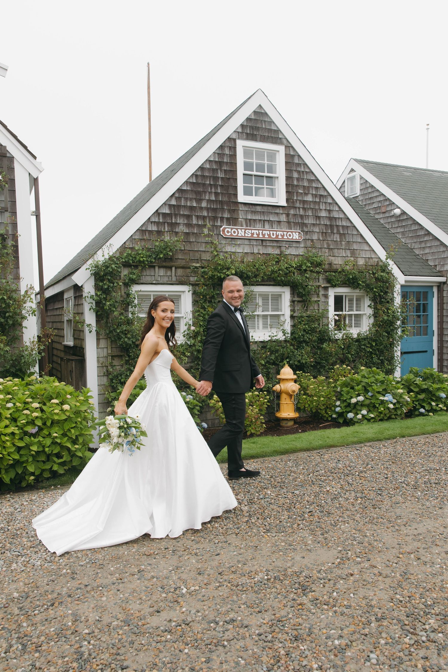 A couple poses near a traditional New England style house with gravel driveway.