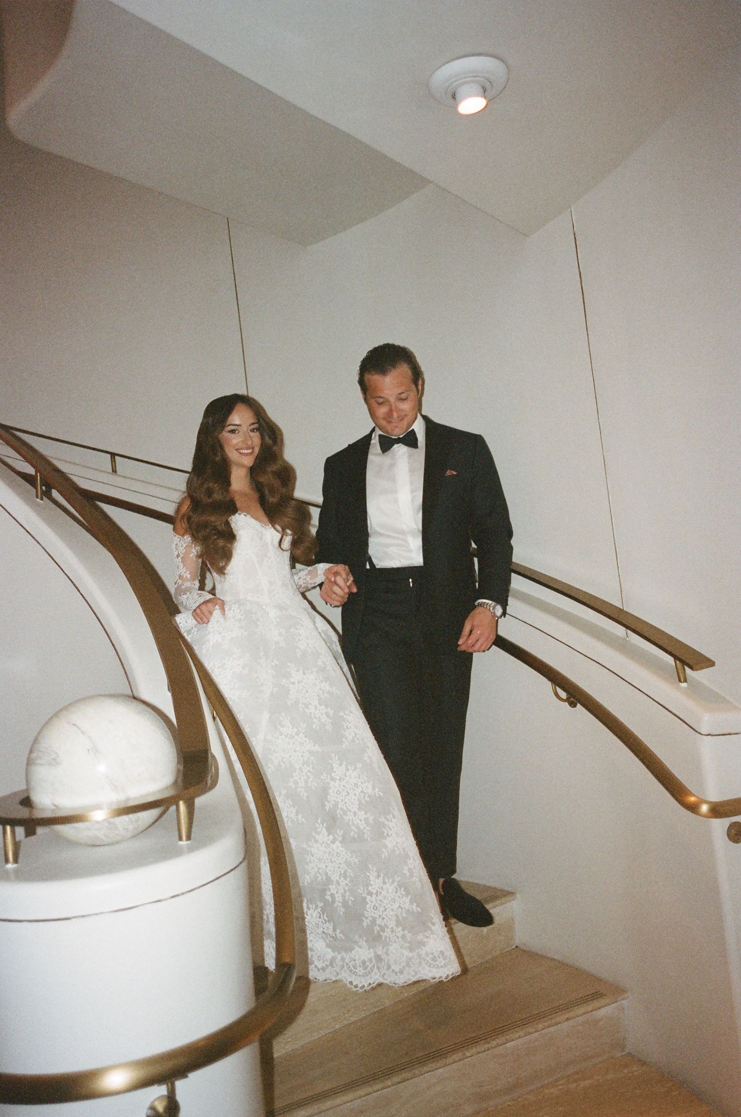 A couple in formal wedding attire descends a curved staircase with a gold handrail in a white interior space.