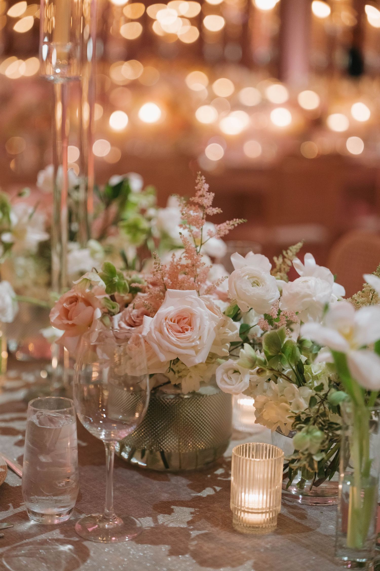 Close-up of wedding table decor featuring blush roses, white flowers, and glowing votives on a neutral tablecloth.