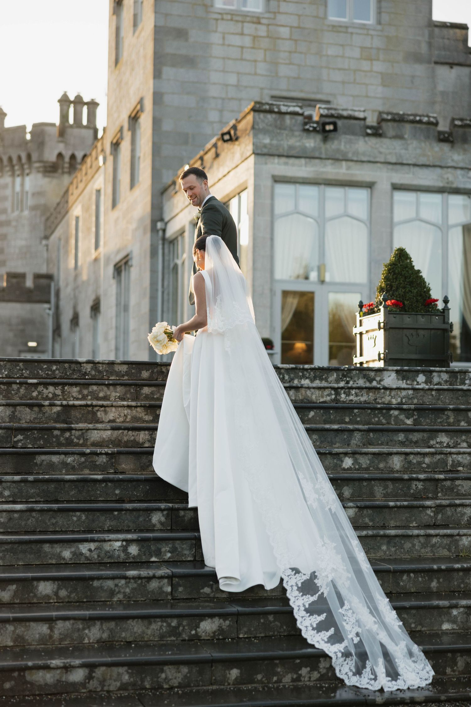 Bride's lace veil trails elegantly down castle steps during wedding photoshoot.