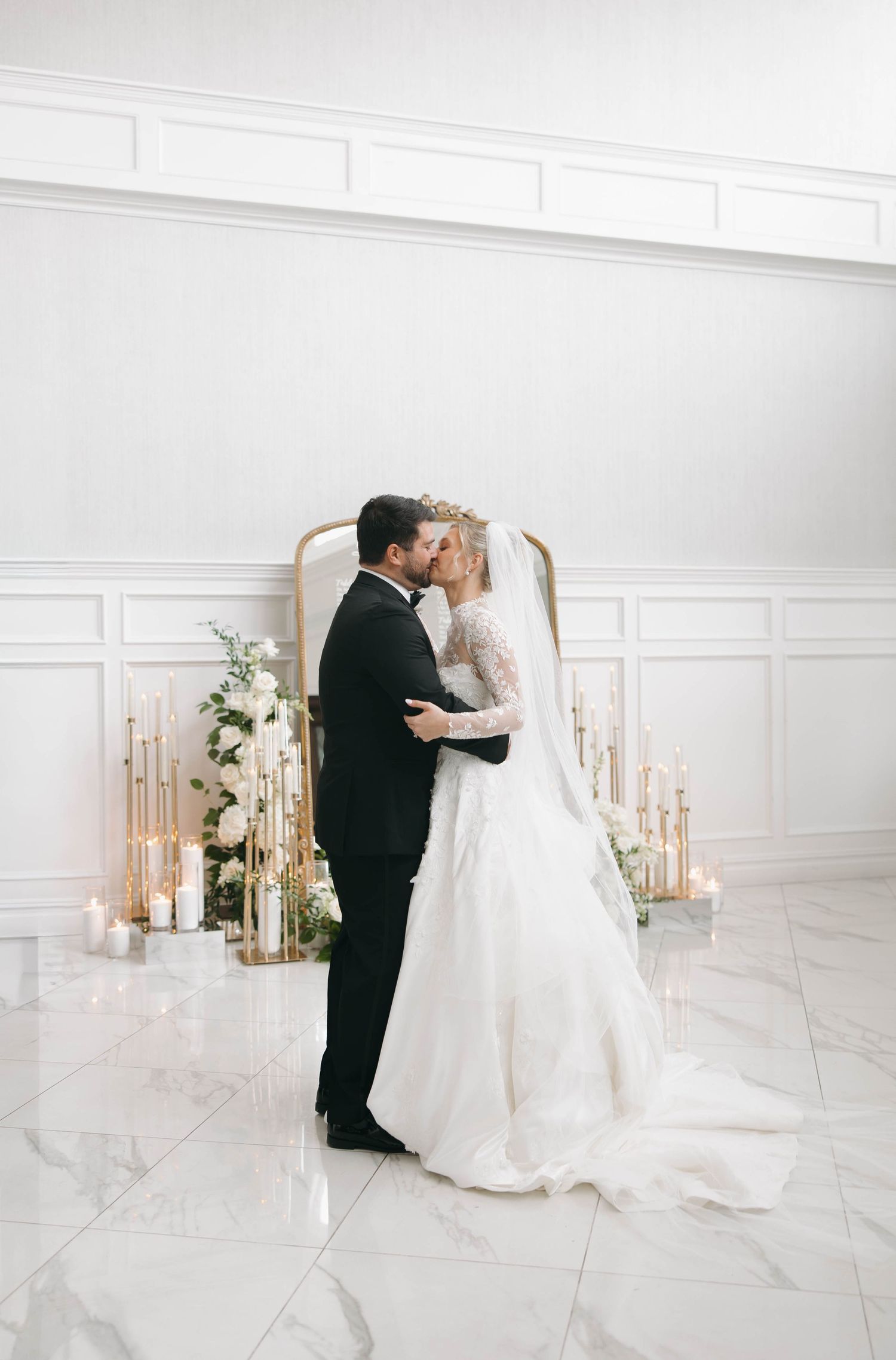Romantic wedding scene in a bright white room with ornate mirror and floral decorations.
